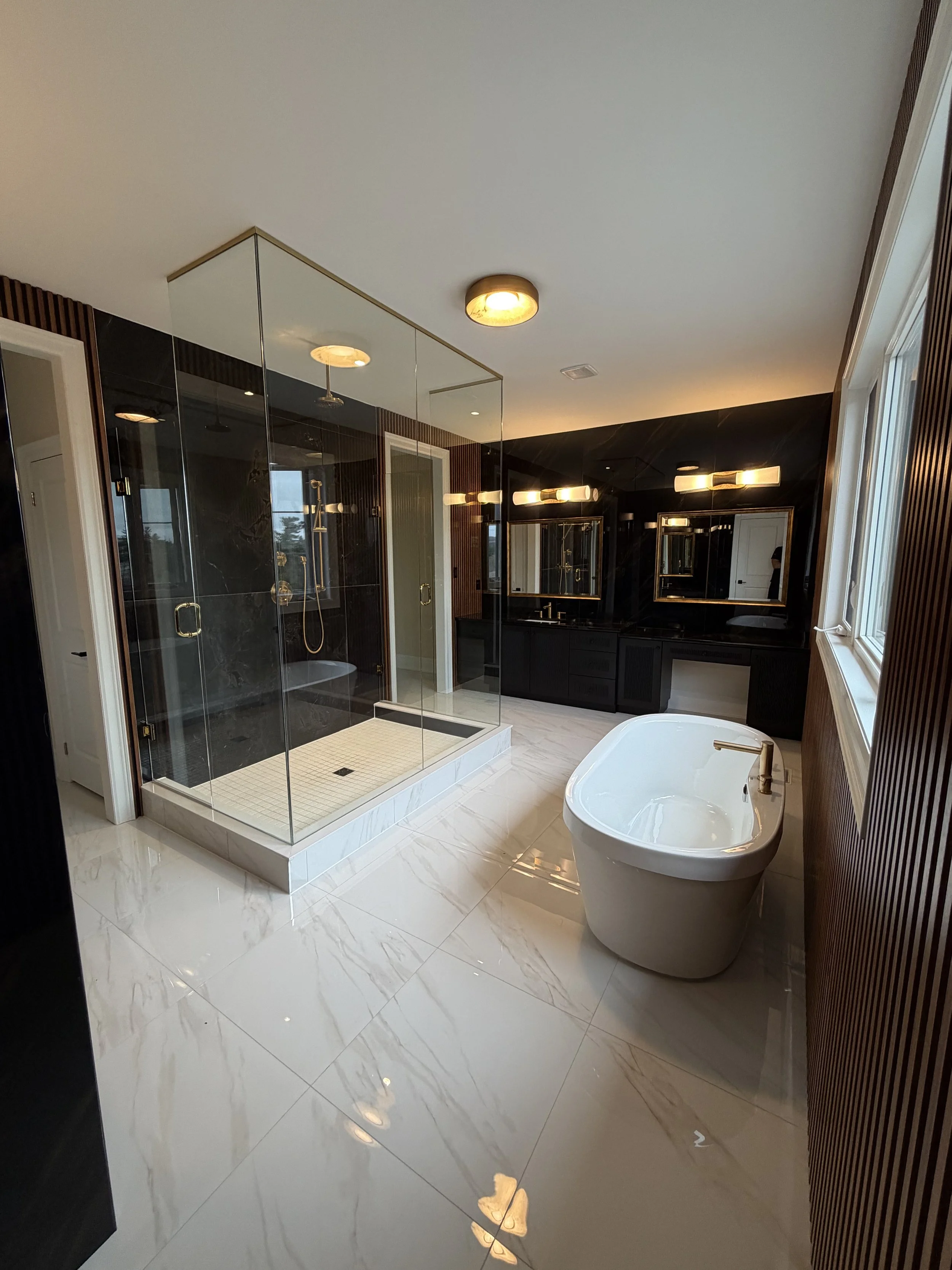Modern bathroom with a walk-in shower with glass doors, a white freestanding bathtub, a large window, dark wall cabinetry, and sleek lighting fixtures.