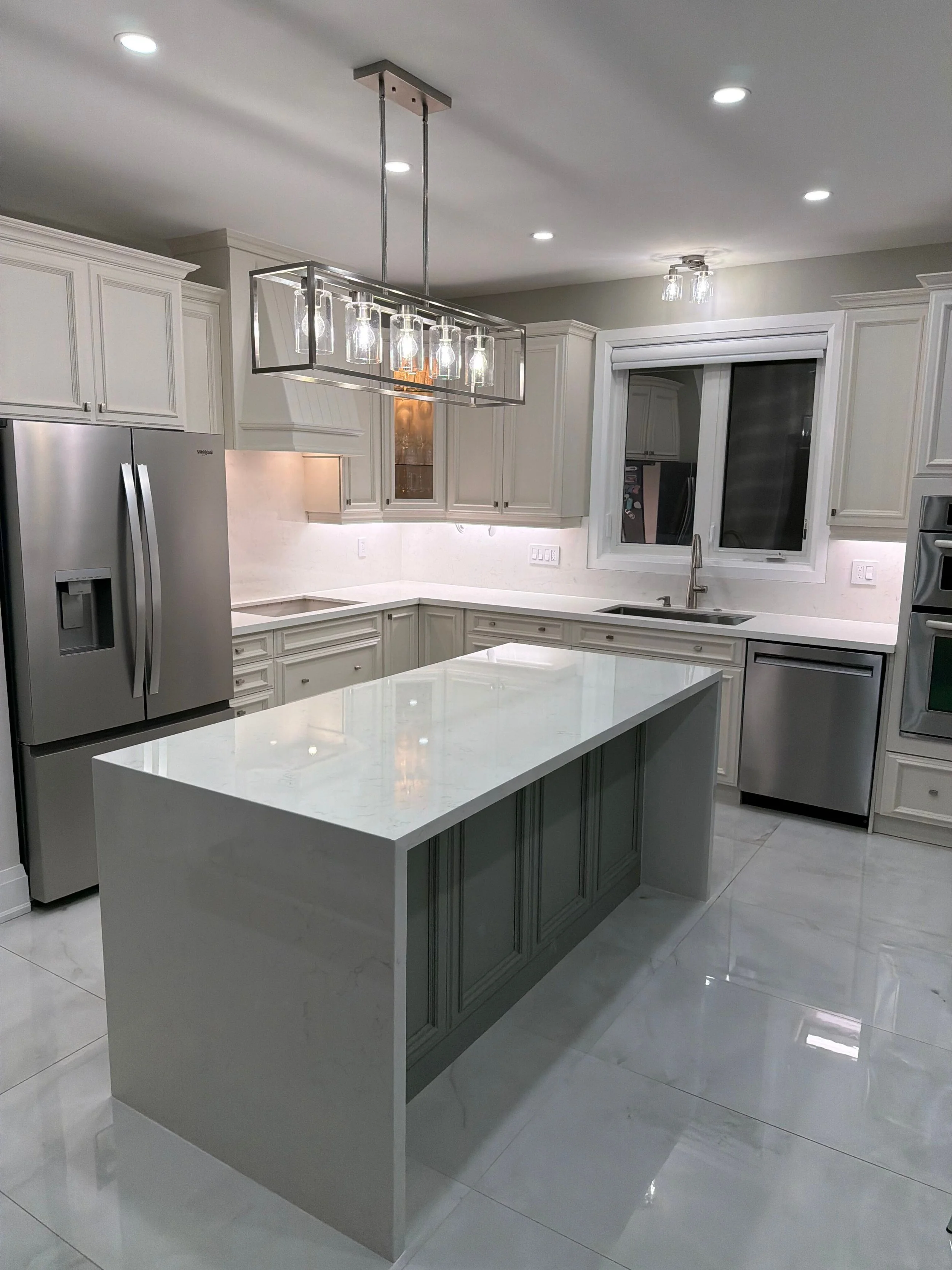 Modern kitchen with white cabinets, marble countertops, a stainless steel refrigerator, and a central island with a white top and light green base, illuminated by ceiling lights and a chandelier.