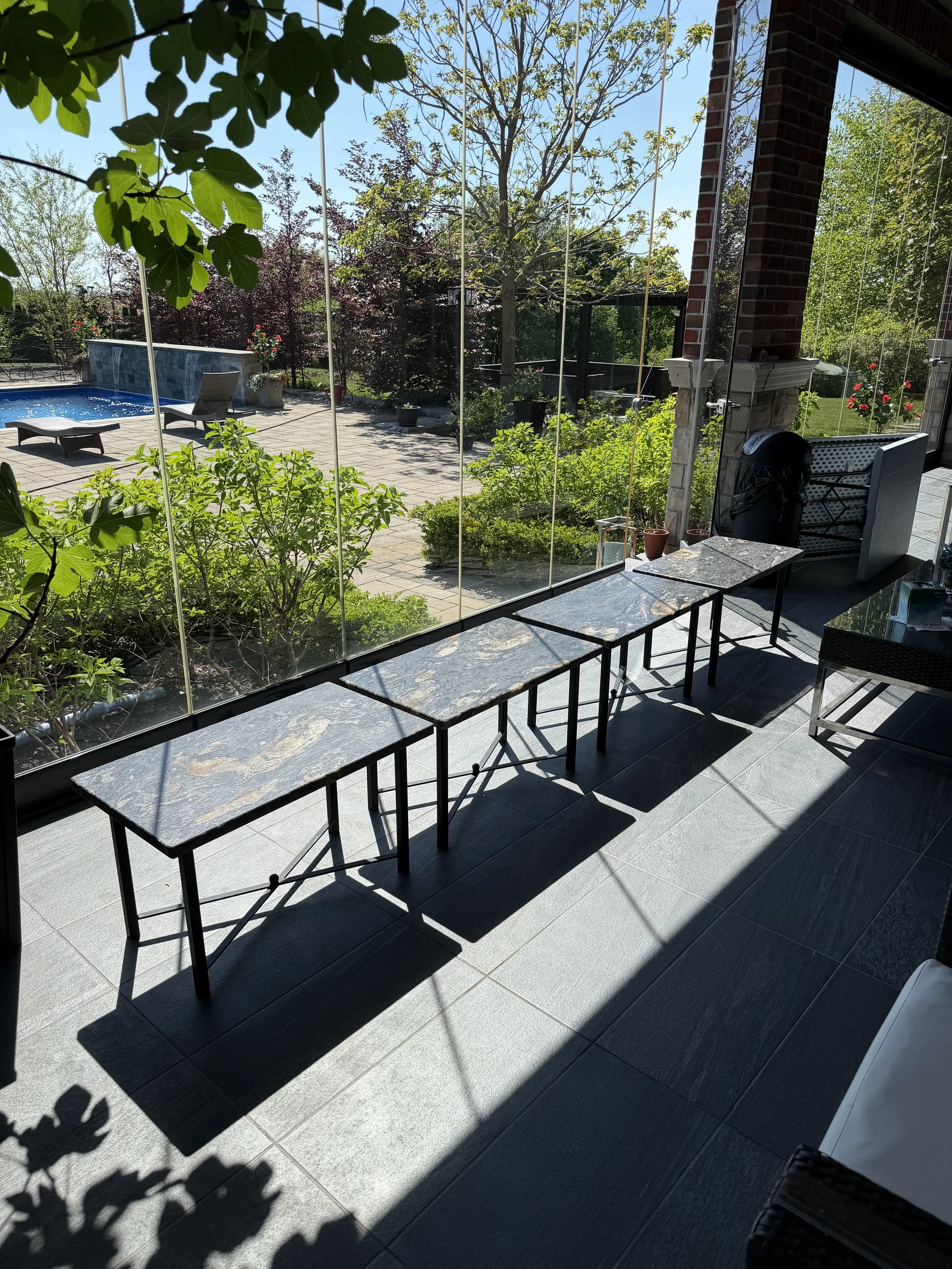 View from inside of a house looking out a glass wall onto a backyard with a swimming pool, trees, and patio furniture. Sunlight casts shadows on the tiled floor.