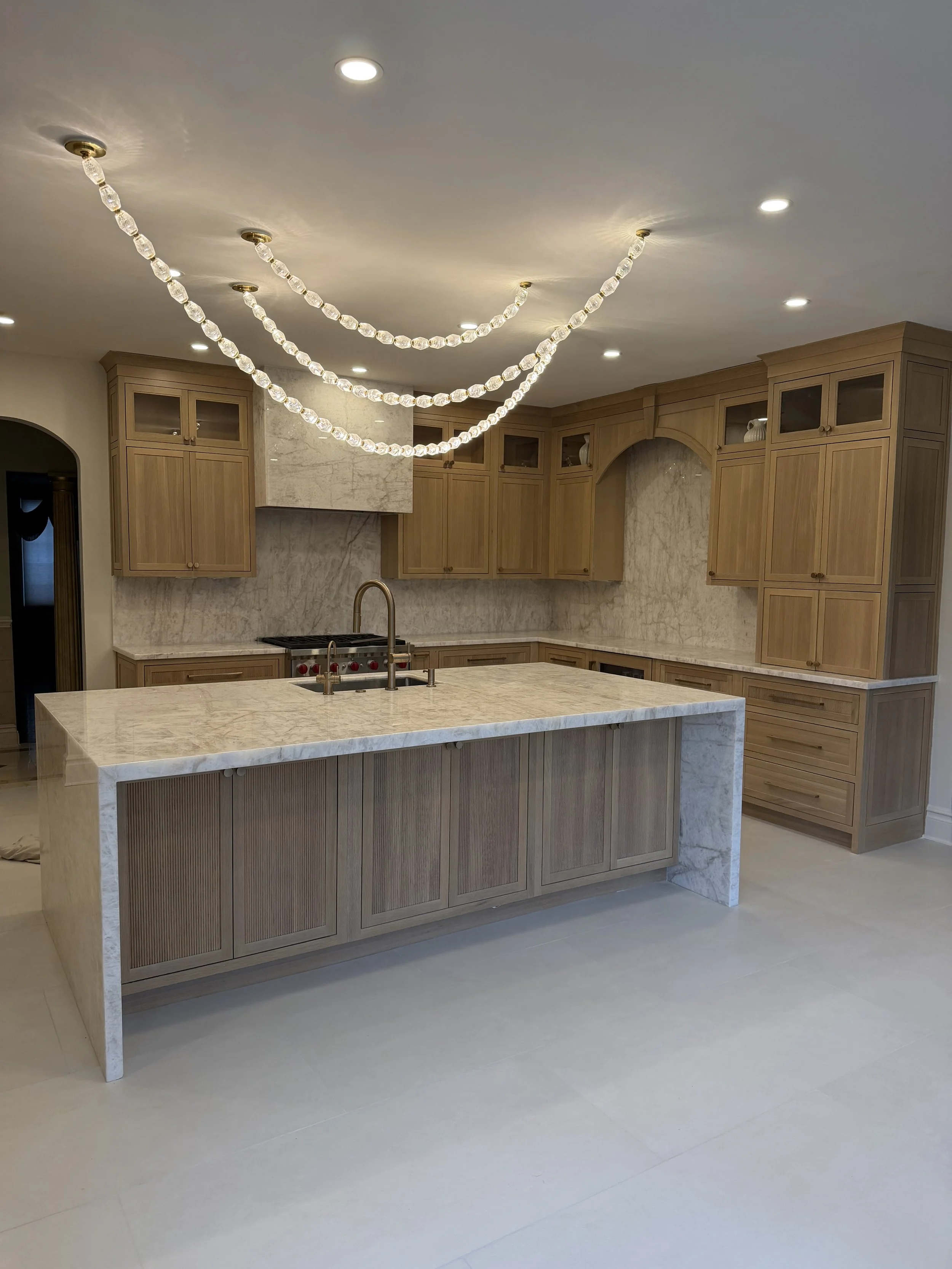 Kitchen with light wood cabinets, marble countertops, and a central island with a marble top. There is a modern chandelier with glass or crystal beads hanging above the island. Recessed lighting is installed in the ceiling.