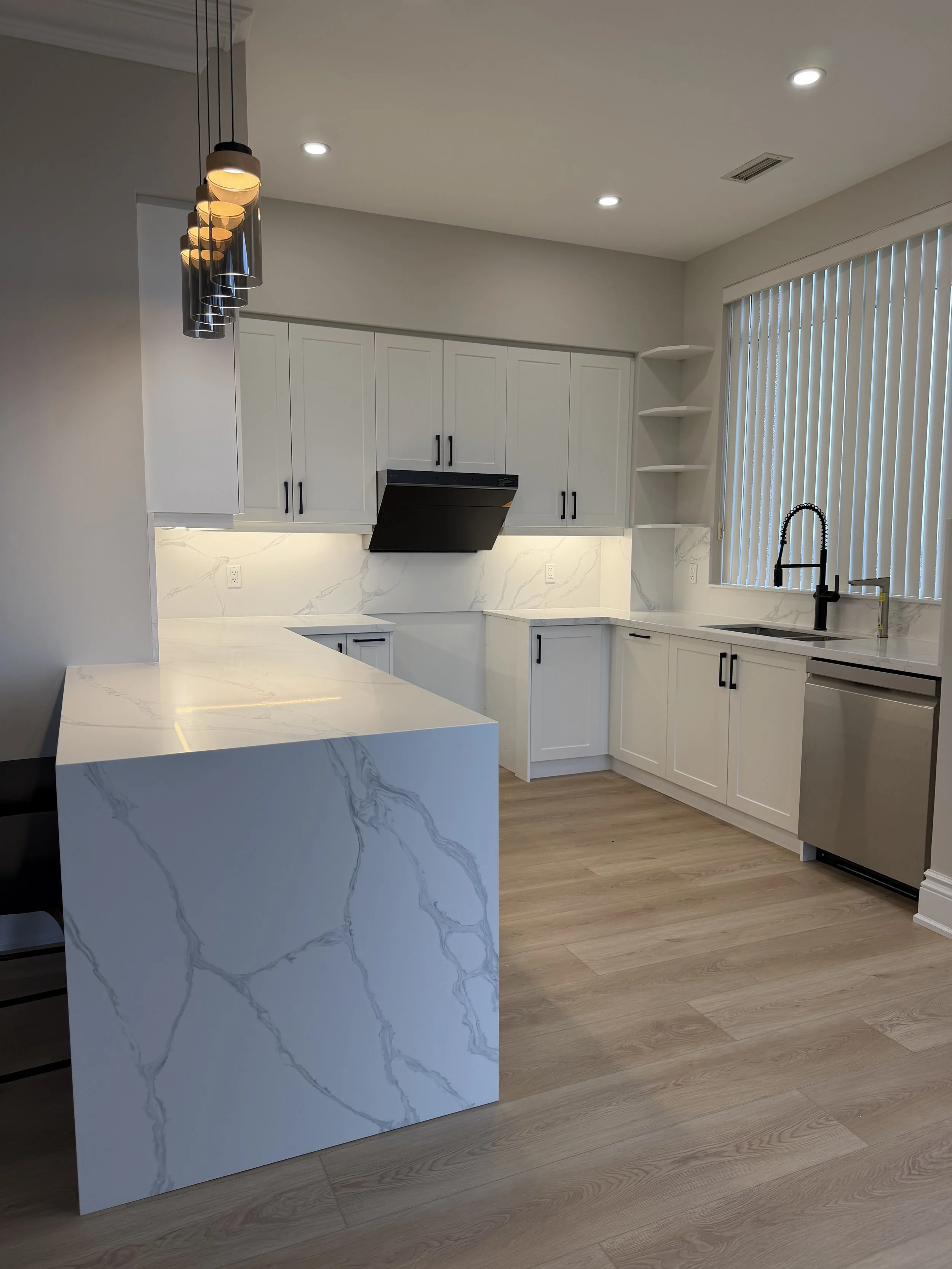 Modern kitchen with white cabinets, marble backsplash, black hardware, black range hood, kitchen sink with black faucet, and large window with vertical blinds.