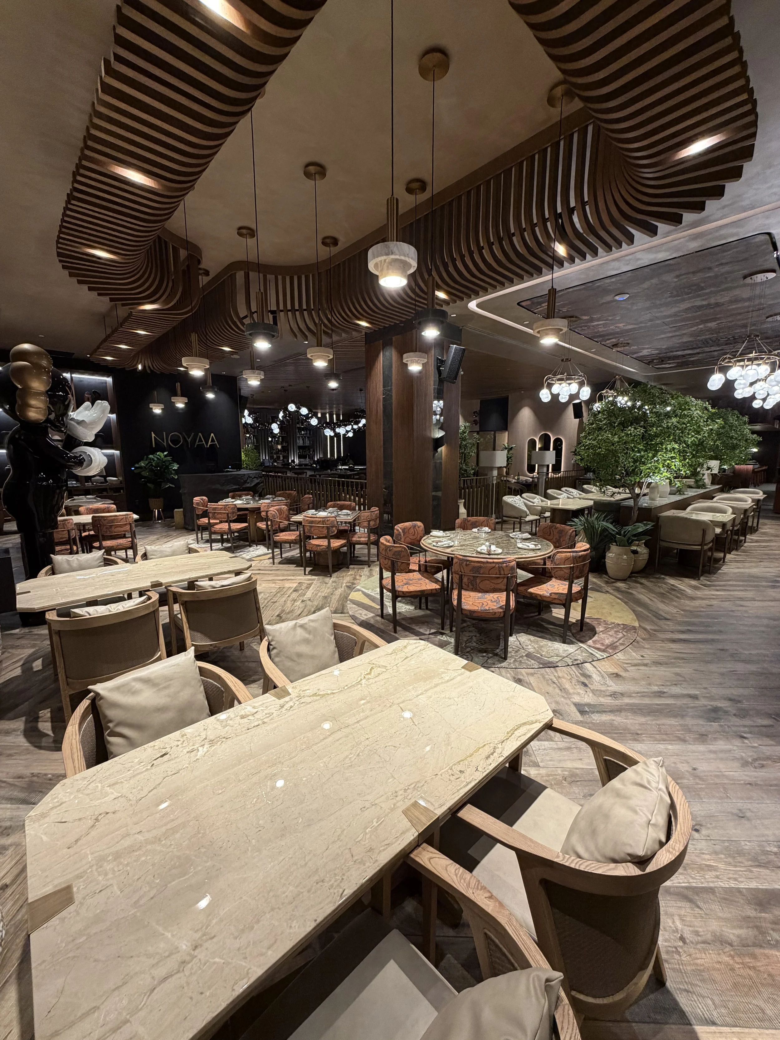 Interior of a modern restaurant with wooden ceiling details, hanging pendant lights, a marble-topped table in the foreground, and various seating arrangements including patterned chairs, couches, and large potted plants.