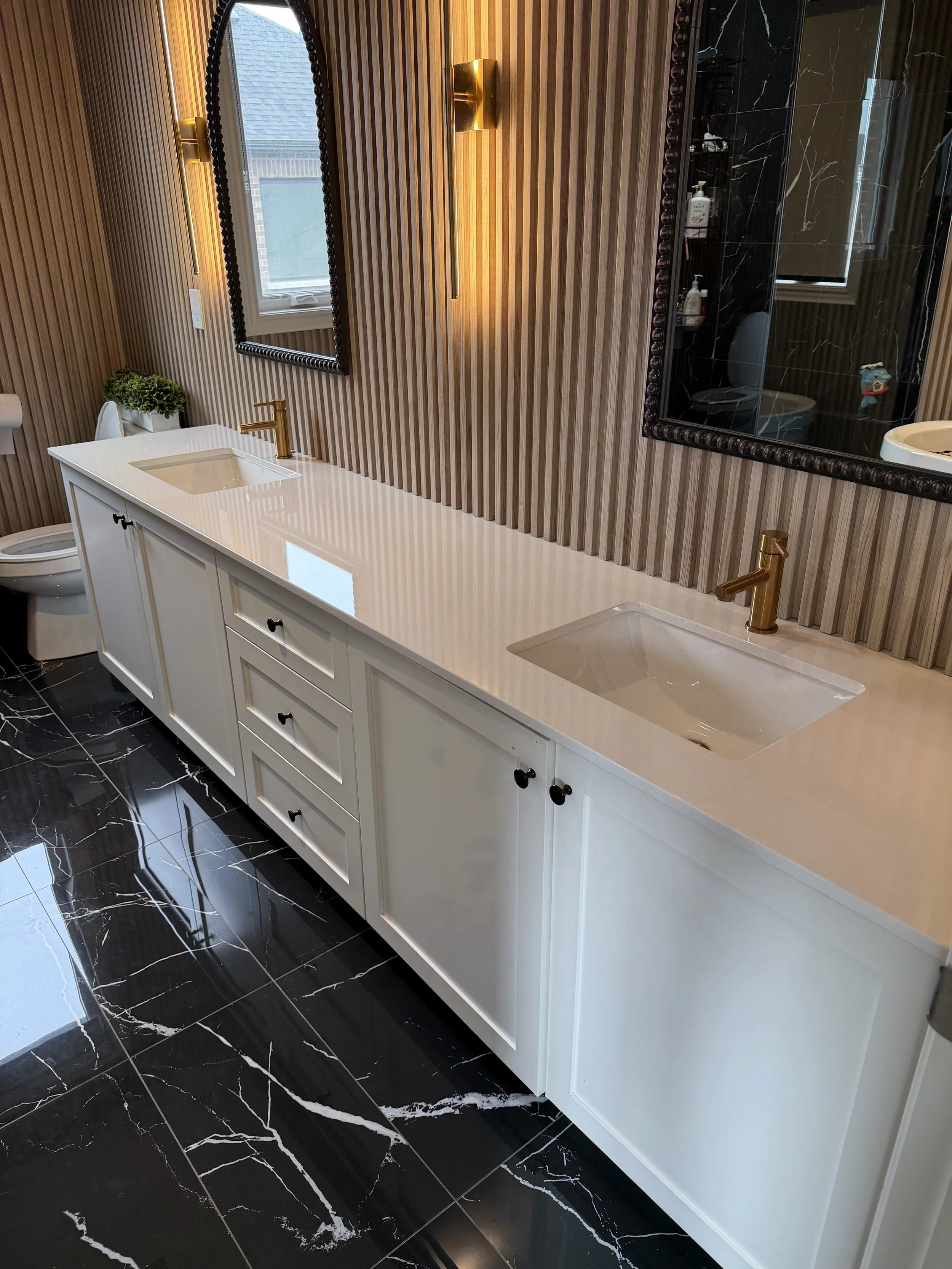 Modern bathroom with black marble floor, white vanity with gold fixtures, and wood-paneled wall with large mirror and window.
