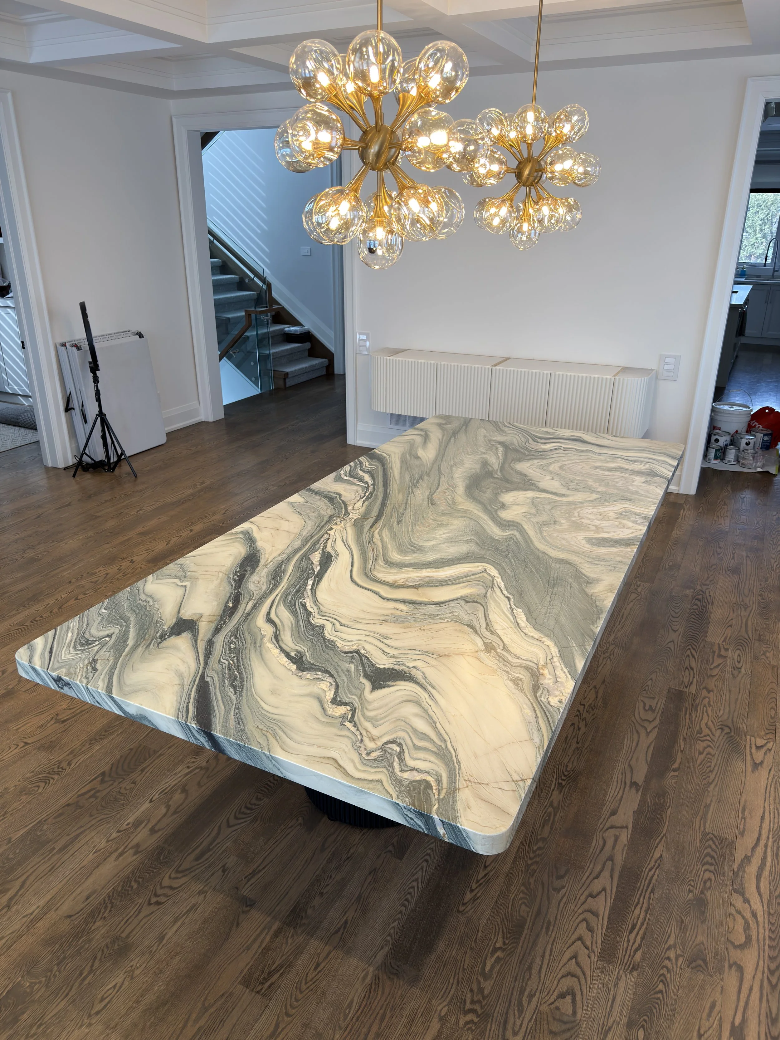 A modern dining room with a large, polished marble table featuring swirling patterns in shades of beige, gray, and black. Two contemporary chandeliers with multiple glass globes hang above the table, and in the background, there is a staircase with a