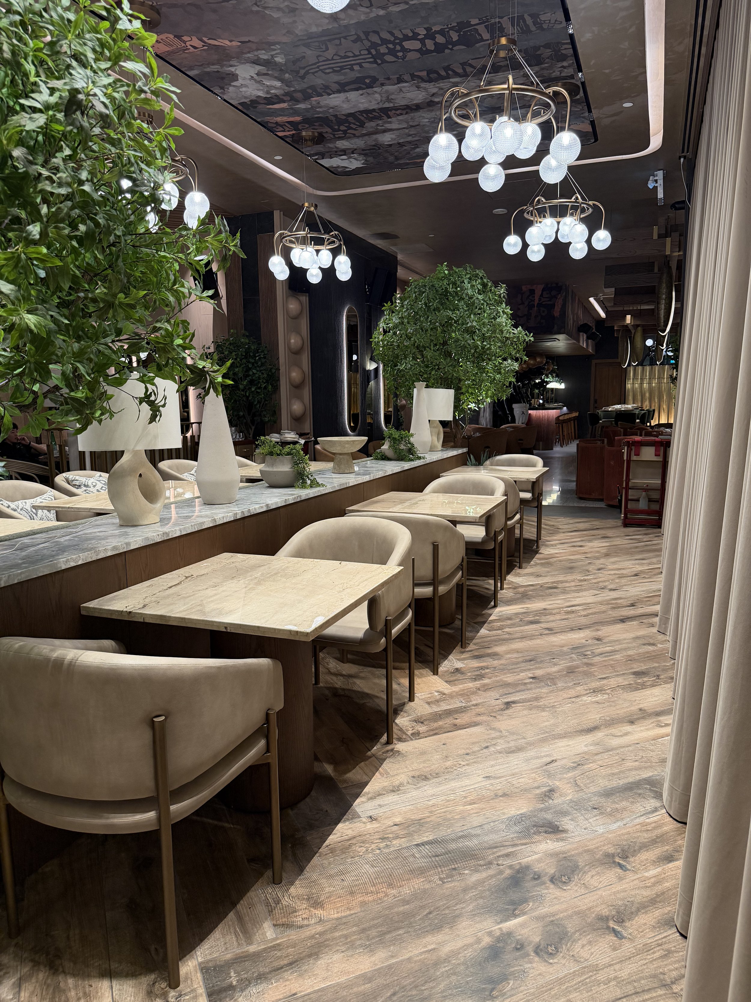 Interior of a modern restaurant with beige chairs around light wood tables, decorative vases, and lush green plants, illuminated by elegant chandeliers with spherical bulbs, and features wood and dark accents.