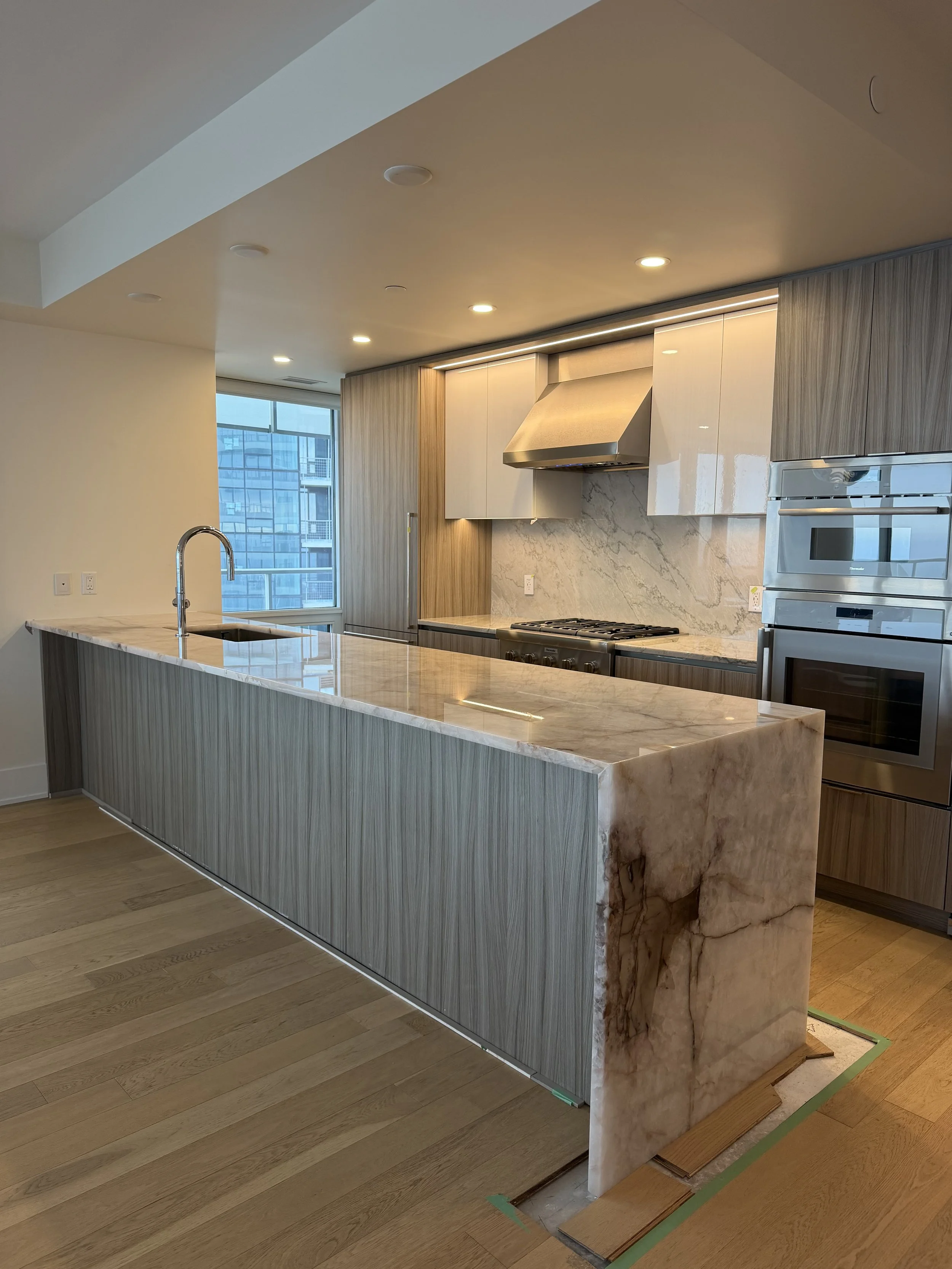 Modern kitchen with marble island, wooden cabinetry, stainless steel range hood, and appliances, with hardwood floors and a large window in the background.