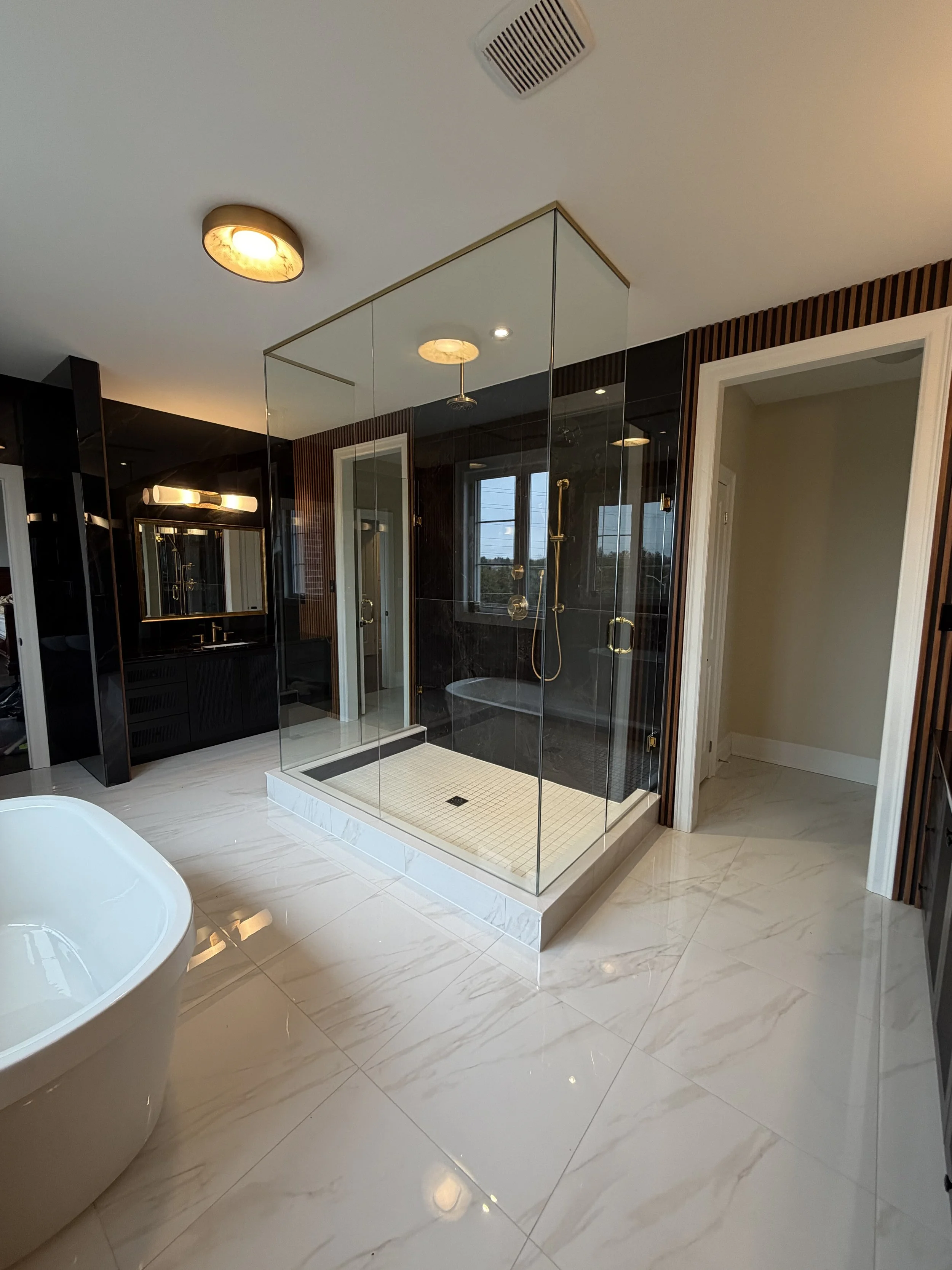 Modern bathroom featuring a glass-enclosed shower with black tiles, a window, a rain showerhead, a white bathtub, a wall-mounted mirror with lighting, and marble flooring.