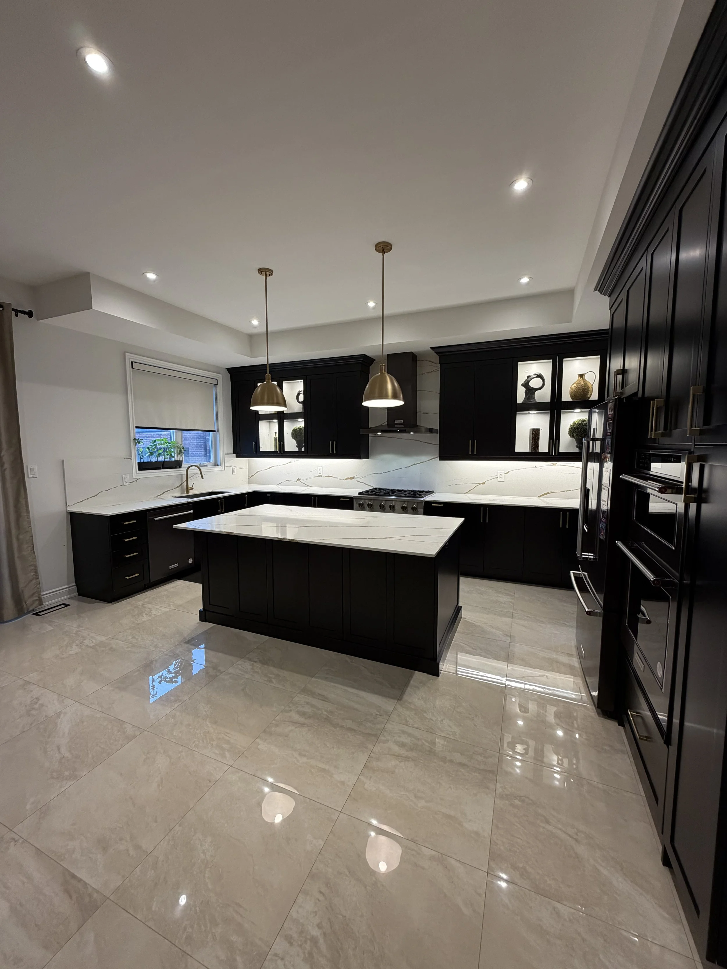 A modern kitchen with black cabinets, a white marble island, a window with a blind, and recessed lighting in the ceiling.