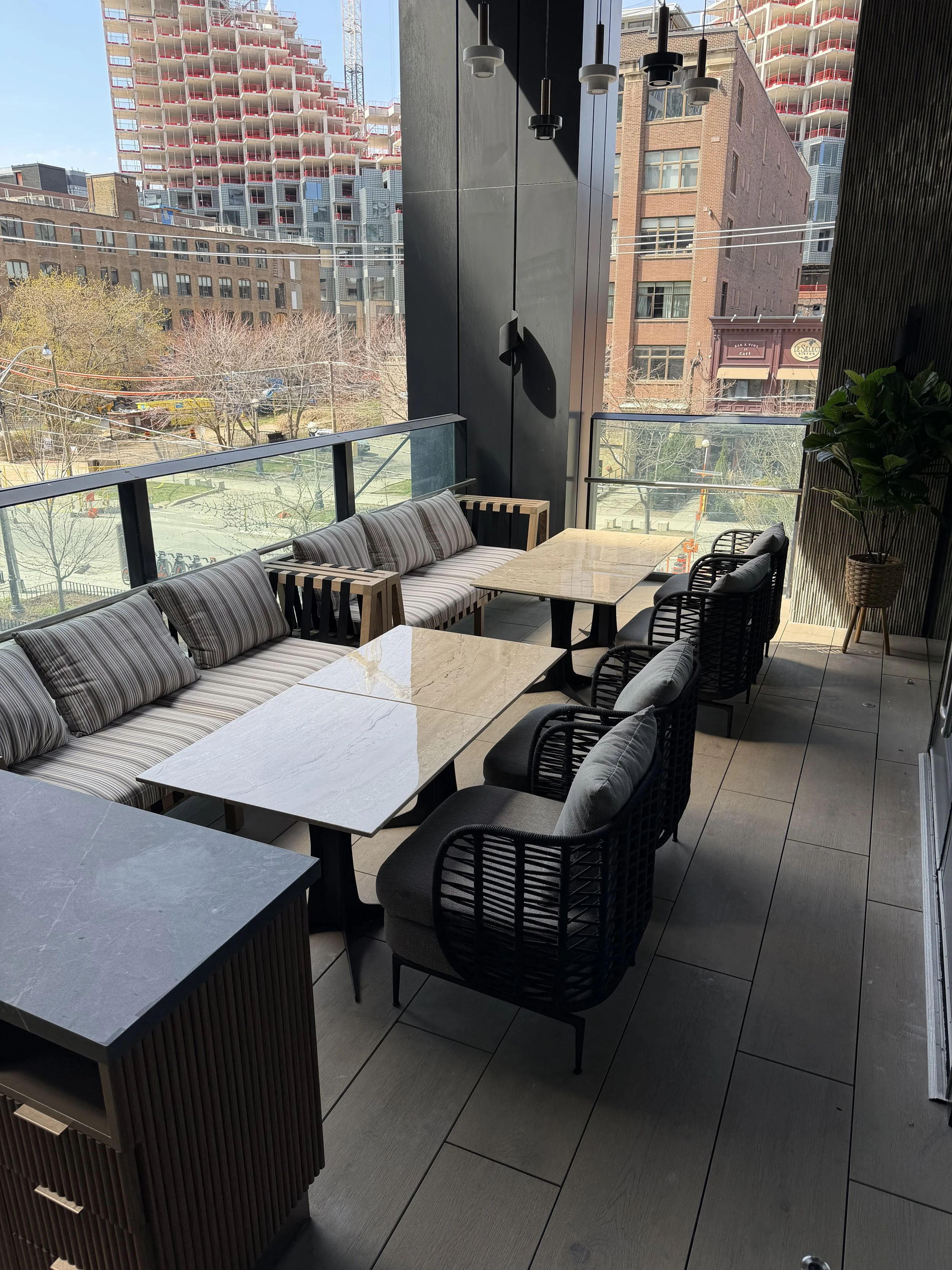 Inside a modern restaurant or cafe, featuring a marble table, cushioned seating, black wicker chairs, and a view of city buildings and trees outside through large glass windows.