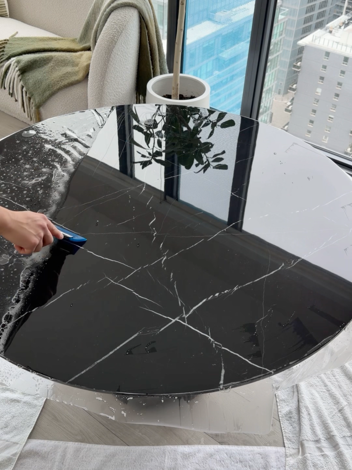 A person is installing TrueGuard Protection film on a marble table. The table is reflective and situated near a large window with a city view, including tall buildings and a plant reflected on its surface.