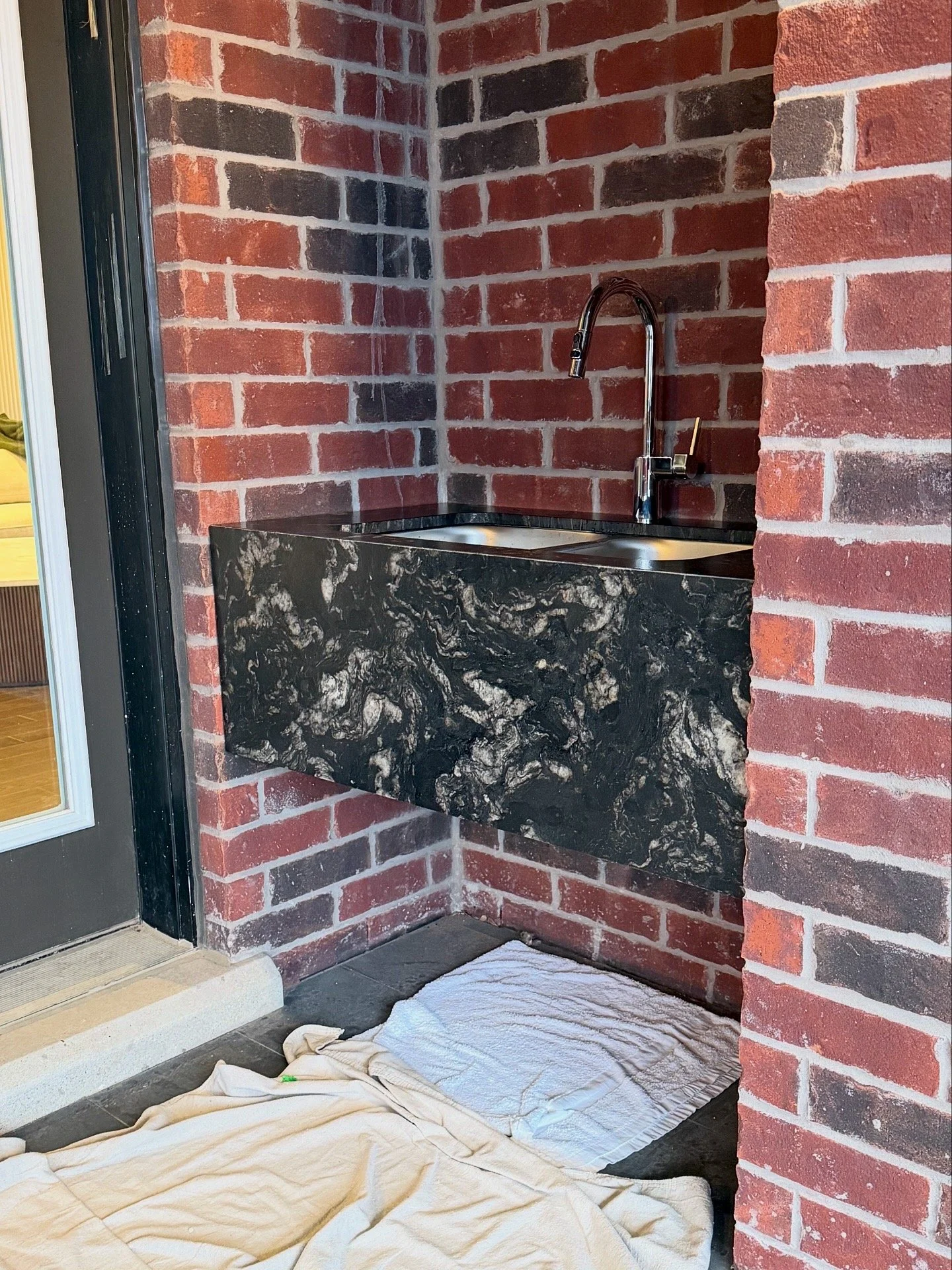 A brick wall with a black marble countertop and a silver faucet. A white cloth is spread on the ground below the counter, and a piece of the cloth hangs off the edge.
