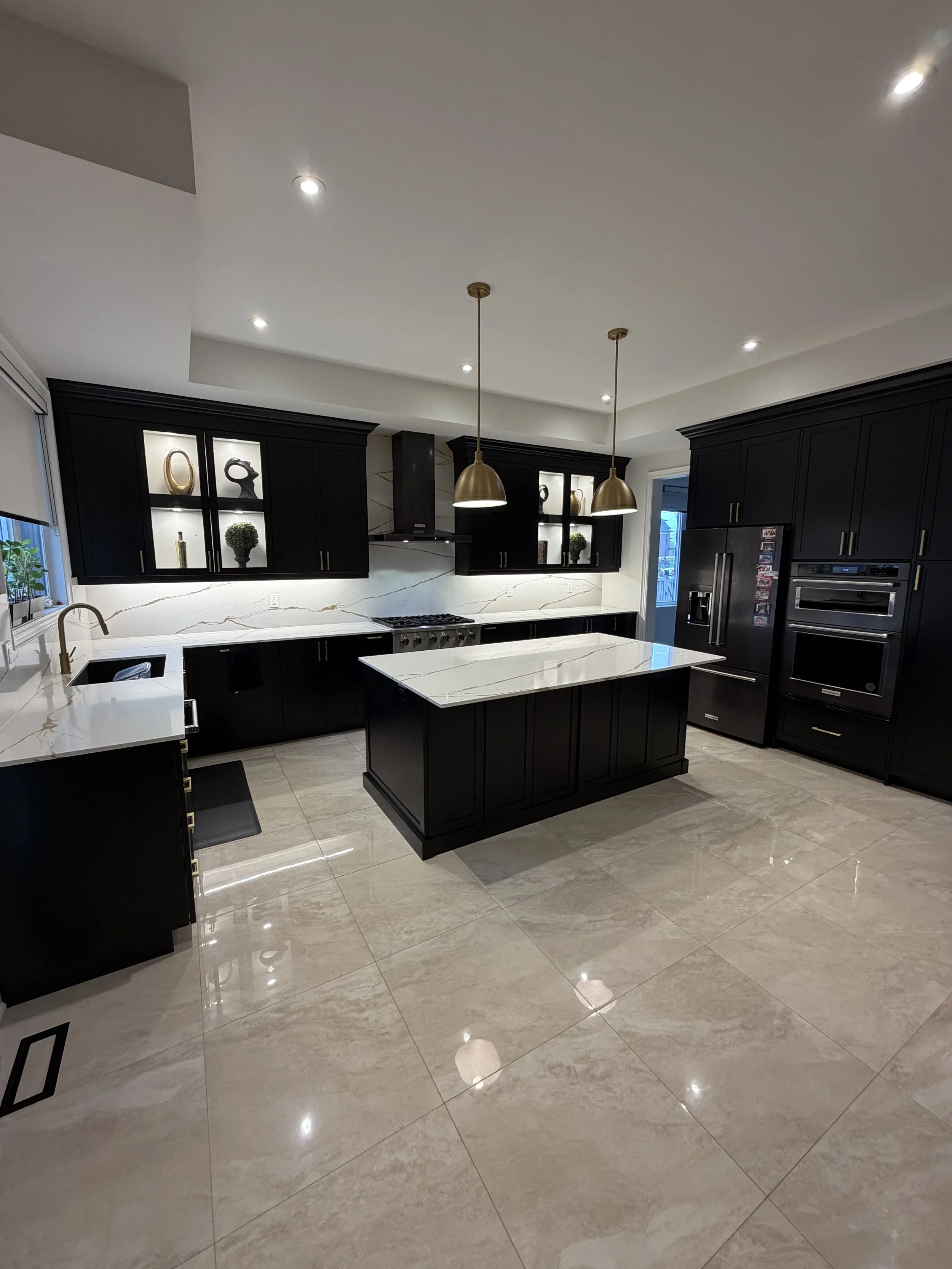 Modern kitchen with black cabinets, white marble countertops, a kitchen island, built-in oven, refrigerator, decorative vases, and pendant lights.