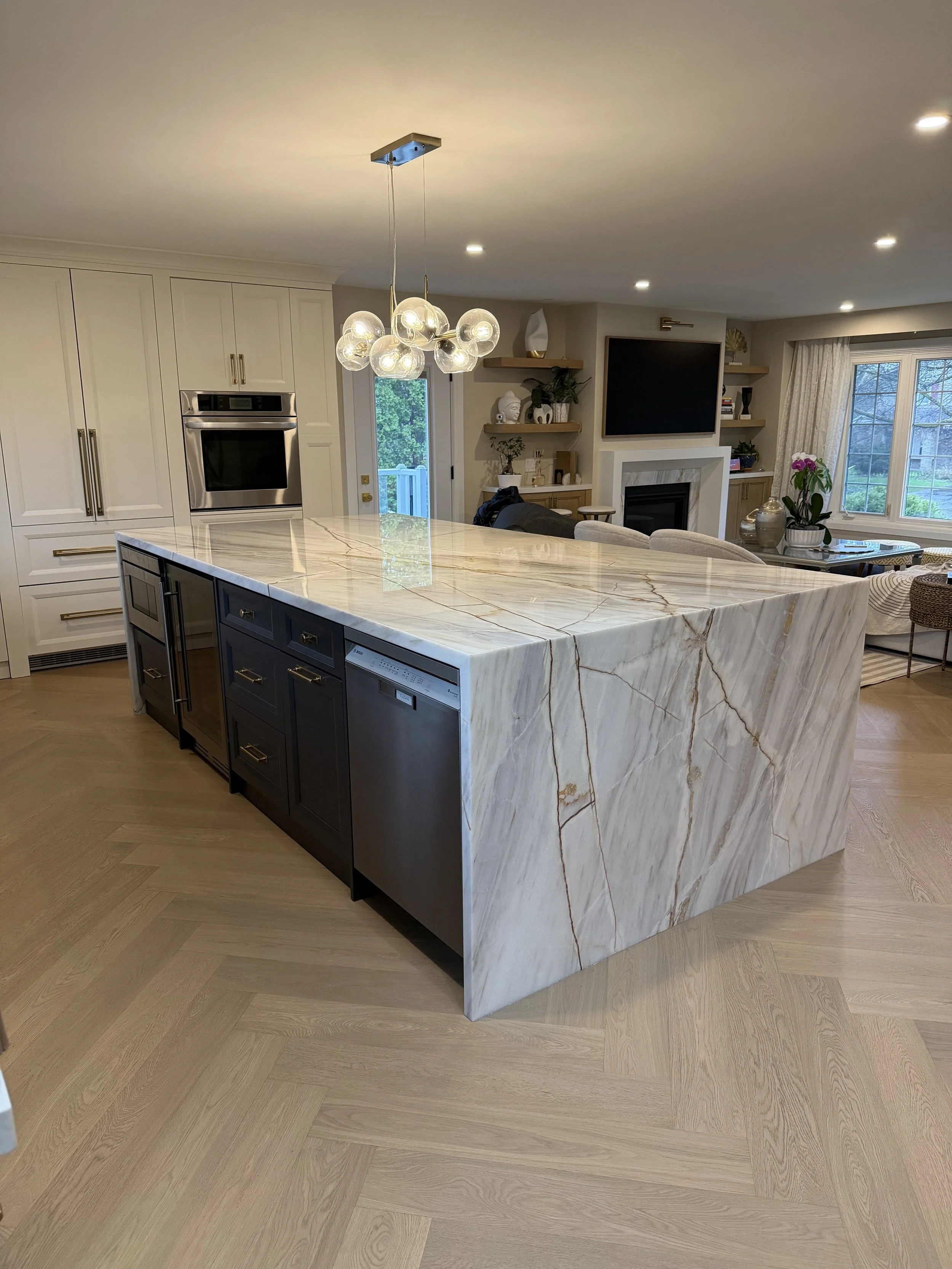 Modern kitchen with marble island, white cabinetry, stainless steel oven, chandelier, and living area with TV and window view.