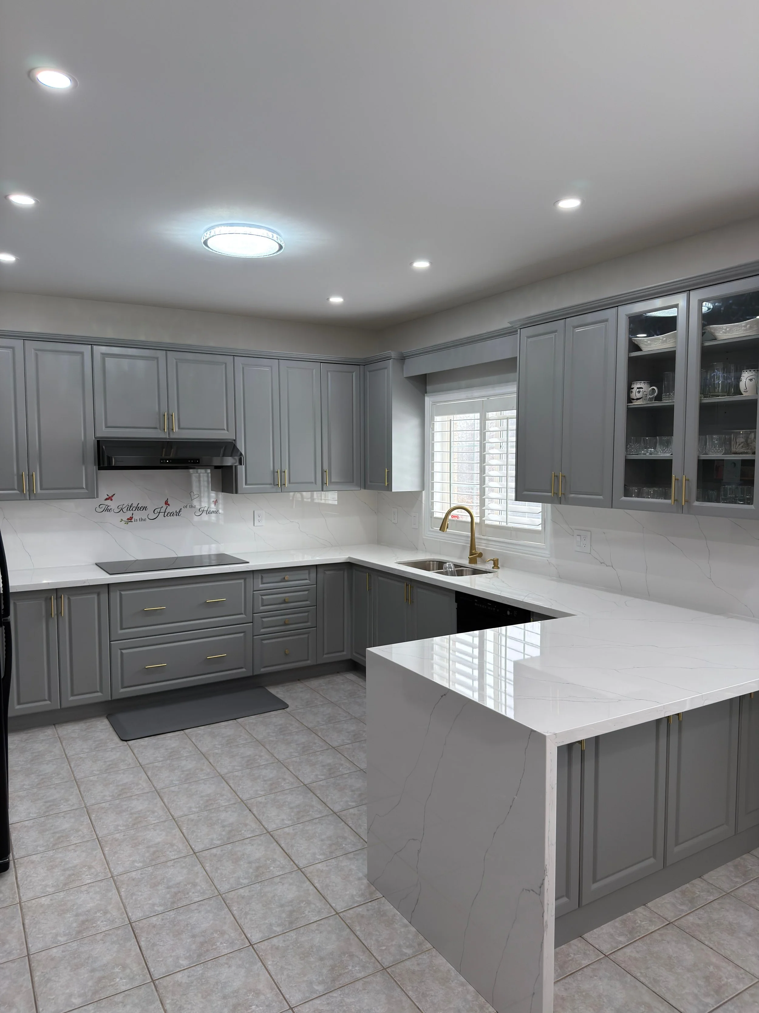 Modern kitchen with gray cabinets, white marble countertops, gold handles, tiled floor, window with shutters, and a black vent hood over the stove.