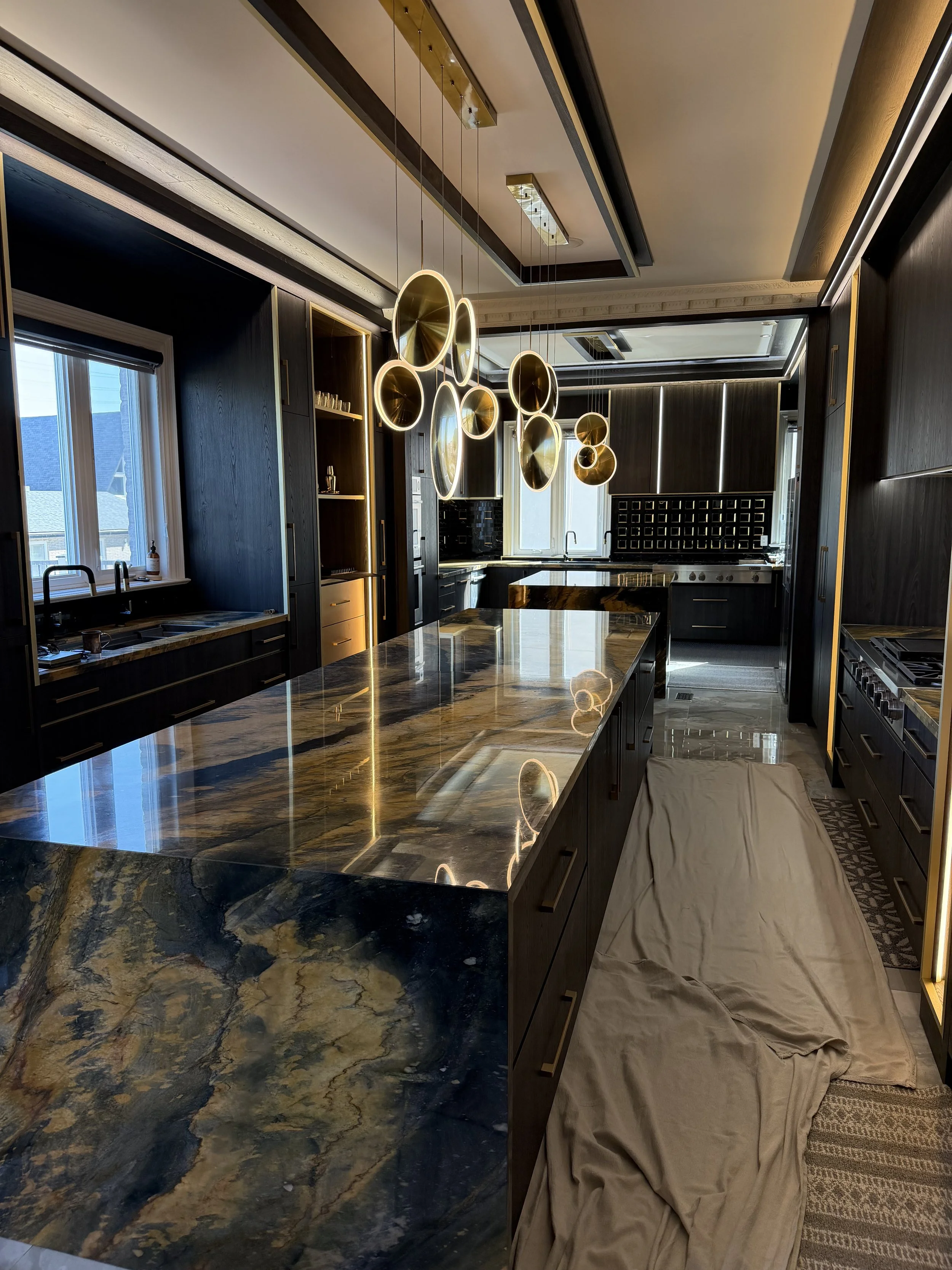 Modern kitchen with dark cabinetry, marble island, large window, decorative pendant lights, and a covered seating area with beige cloth draped over it.