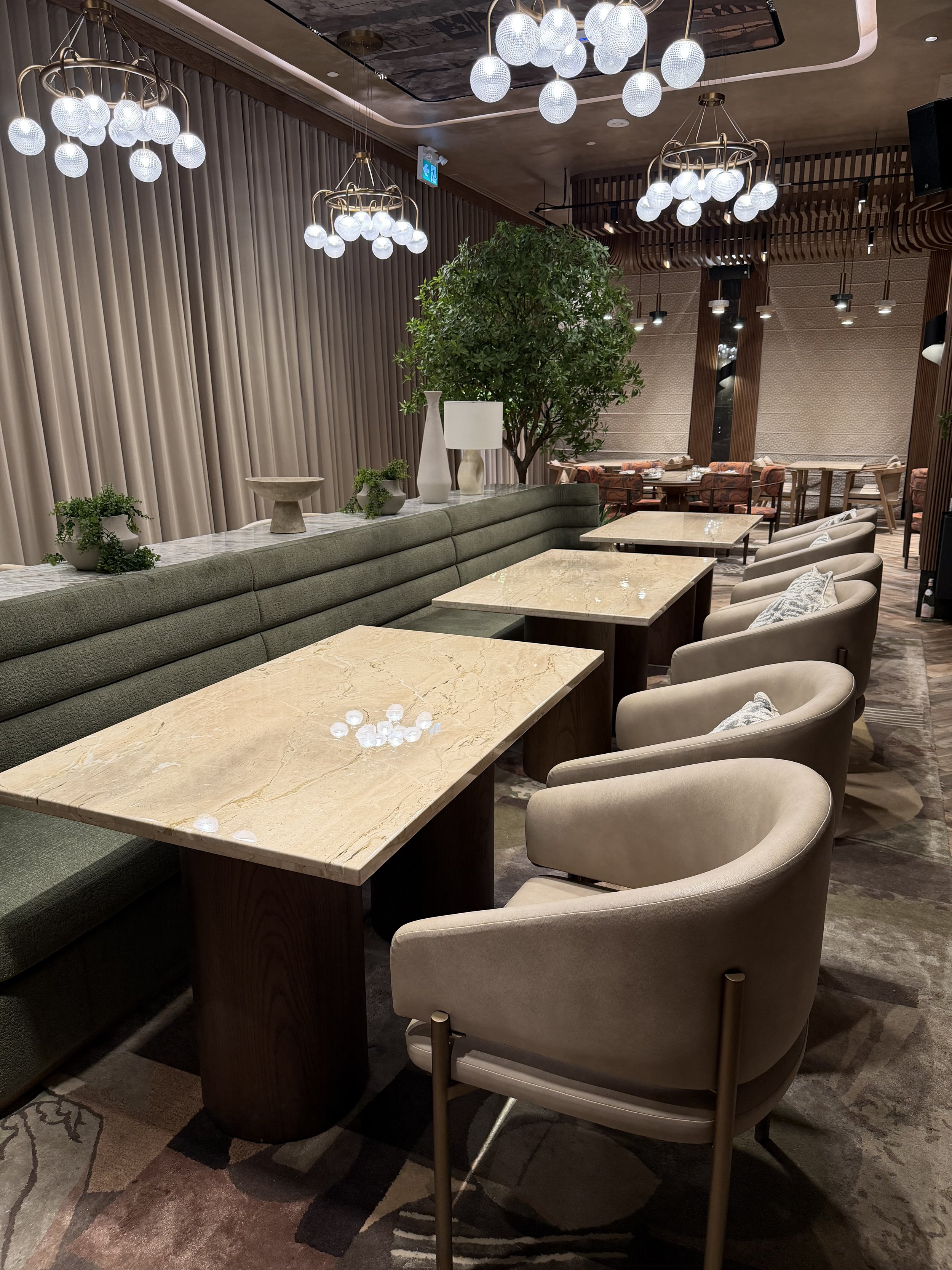 The image shows a modern, upscale lounge or restaurant interior with beige chairs, a long green upholstered sofa, and a marble-topped table in the foreground. Potted plants and decorative vases are placed along the sofa. The ceiling features stylish 