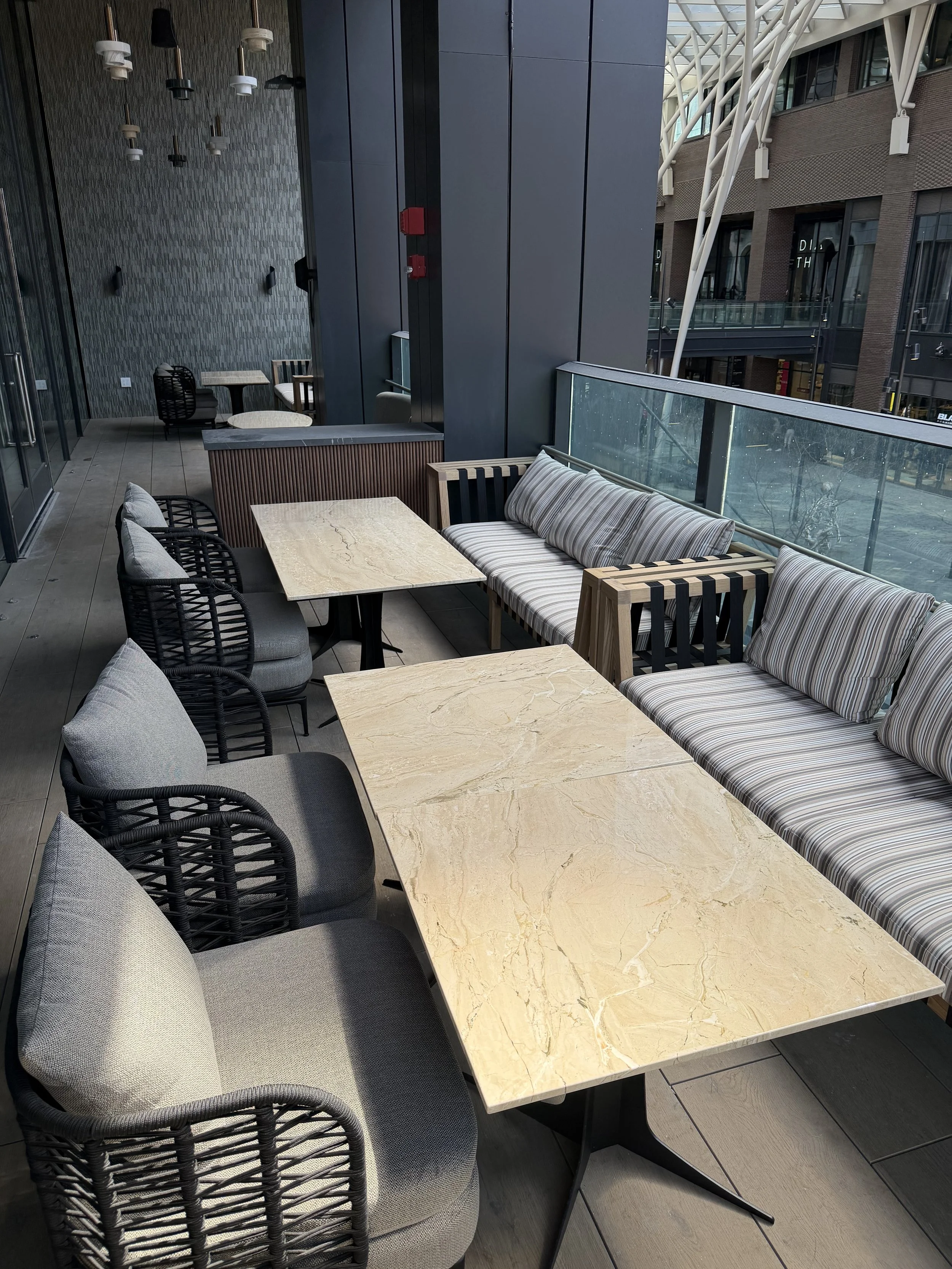 A balcony with beige marble tables and mixed chairs, including woven black chairs and cushioned seats, overlooking a modern city street with buildings and glass railing.