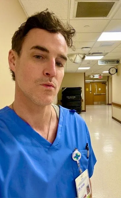 A male healthcare worker wearing blue scrubs and a hospital ID badge taking a selfie in a hospital corridor. The corridor has a clock, ceiling lights, and a door at the end.