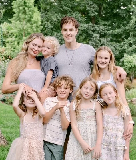 A family of seven standing outdoors in front of green trees, smiling and posing for a photo.