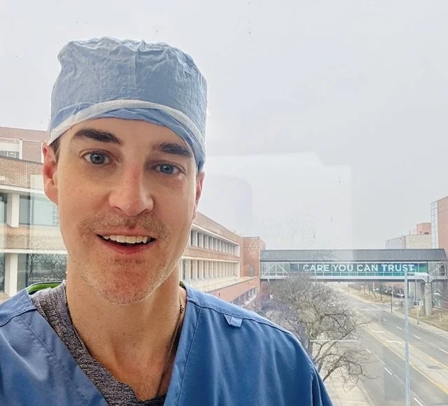 A male healthcare worker in blue scrubs and a surgical cap smiling in a clinical setting with a city street in the background.