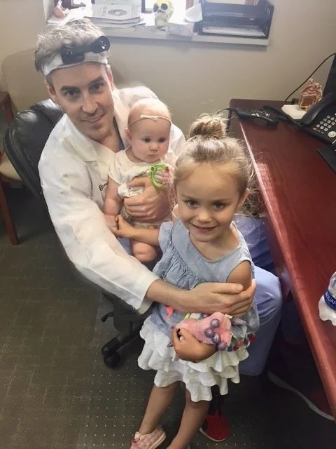 Doctor in white coat and headlamp holding a baby girl with a pacifier, smiling as a young girl in summer dress hugs him in an office setting.