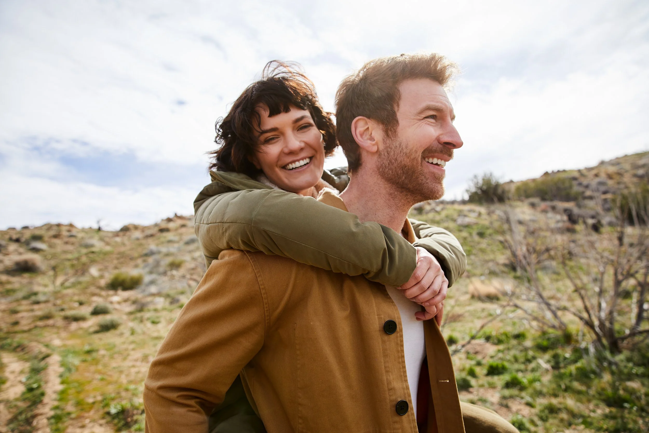 A smiling woman is giving a piggyback ride on a man in a field with sparse vegetation under a cloudy sky.