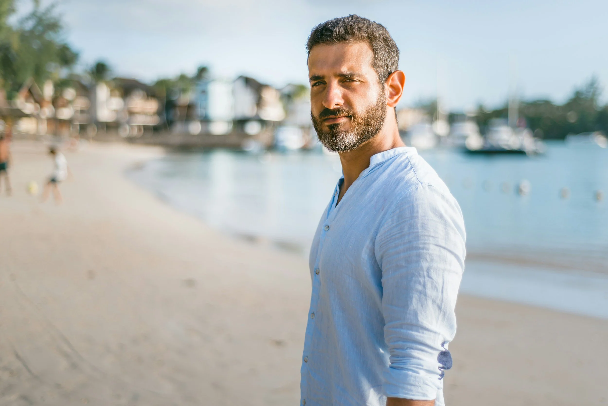 A man with a beard and short dark hair wearing a white shirt with rolled-up sleeves standing on a beach, with houses and boats in the background.