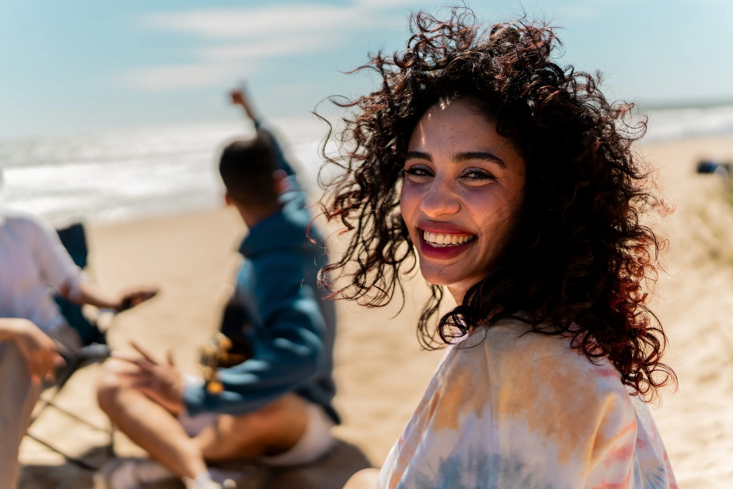 Smiling woman with curly hair at the beach, with people in the background sitting on the sand and waves in the ocean.