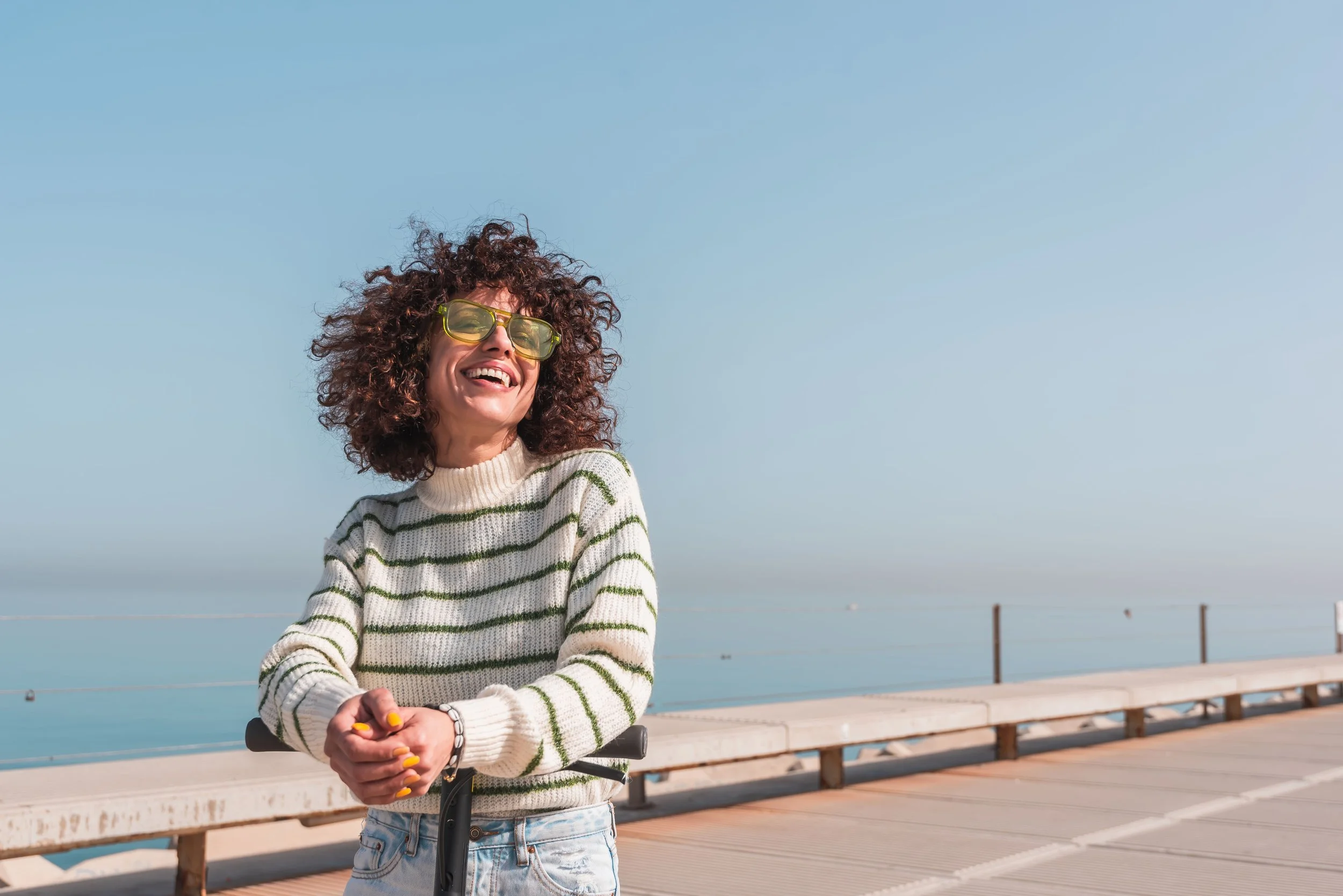 Woman with curly hair wearing yellow sunglasses, a striped sweater, and jeans, smiling and standing on a boardwalk by the water on a bright, clear day.