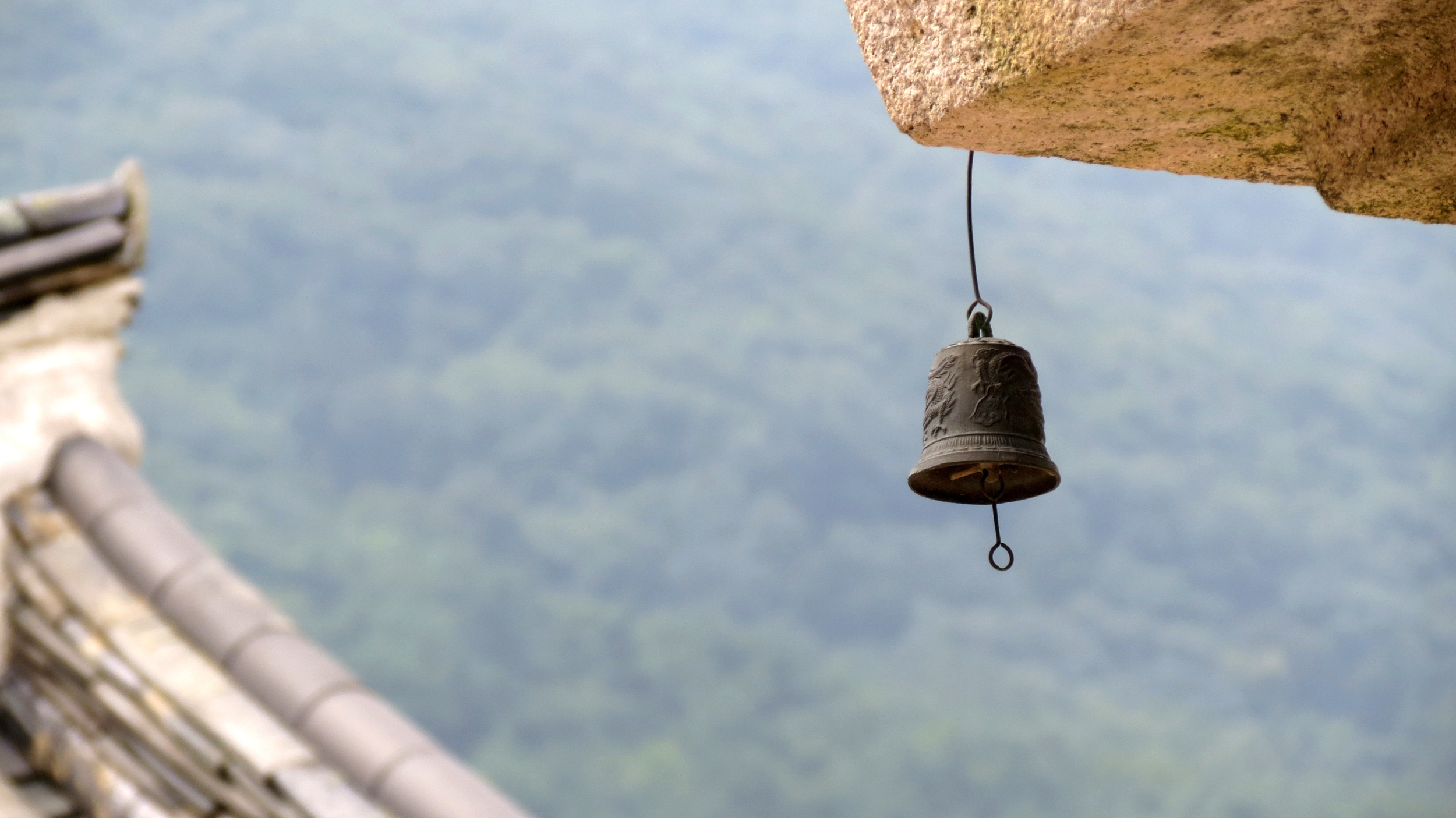 A metal bell hanging from a wire attached to a large rock, with a blurred traditional building with bamboo roof to the left, and a mountain landscape in the background.