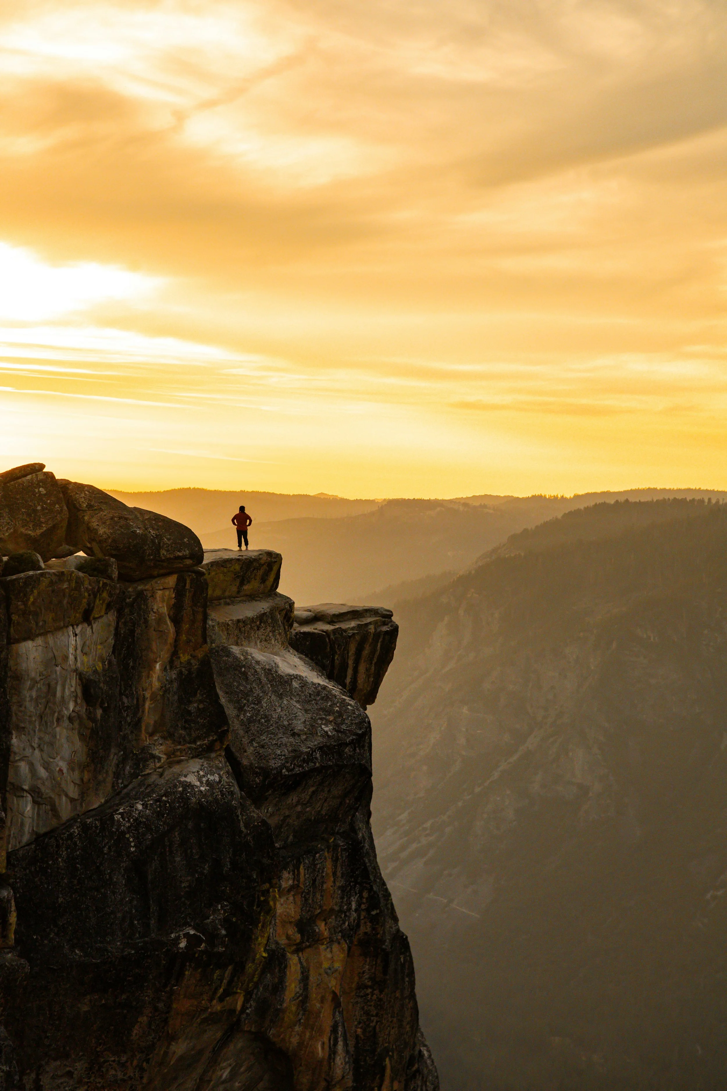 A person standing on the edge of a rocky cliff overlooking a valley at sunset.