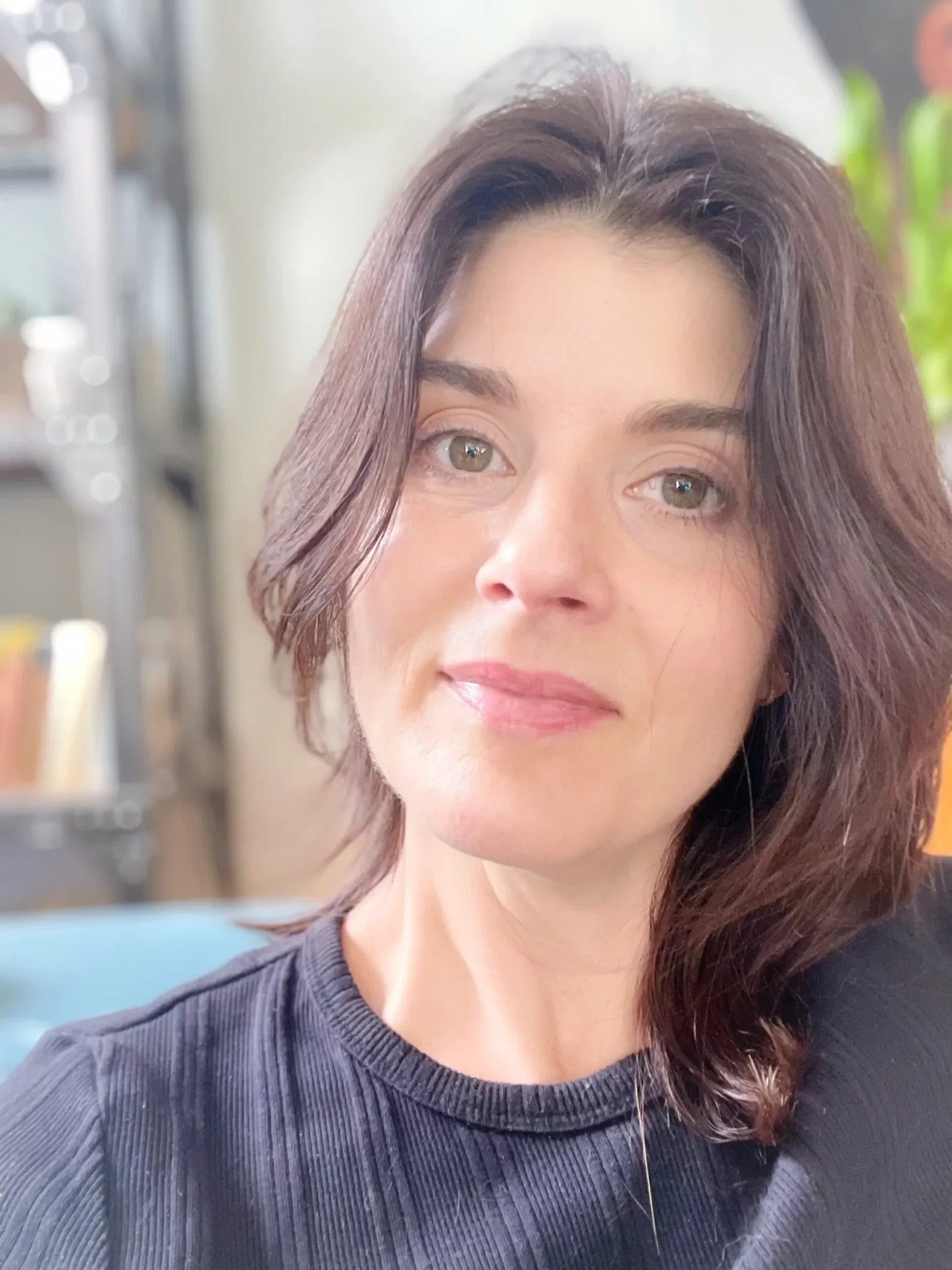 Close-up of a woman with shoulder-length brown hair, light skin, and green eyes, wearing a dark ribbed shirt, smiling softly.