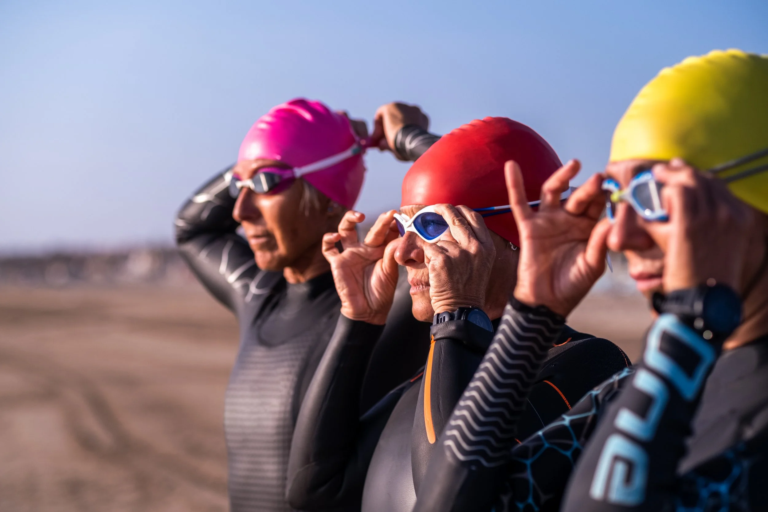 Three athletes wearing black wetsuits and colored swim caps preparing for a race, adjusting their goggles.