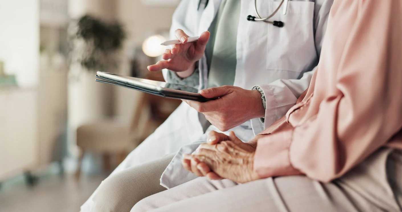 A healthcare professional in a white coat with a stethoscope is talking to an elderly person wearing a pink jacket, in a medical setting.