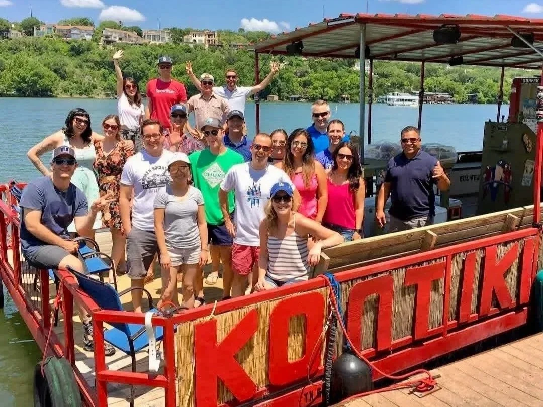 Group of people posing on a boat docked on a river, with a scenic green hillside and houses in the background, under a partly cloudy sky.
