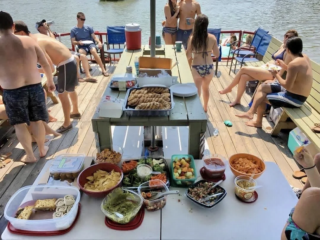 A group of people on a wooden dock at the water's edge enjoying a boat party with food and drinks, some sitting on benches, others standing, with a table of snacks and a grill at the center.