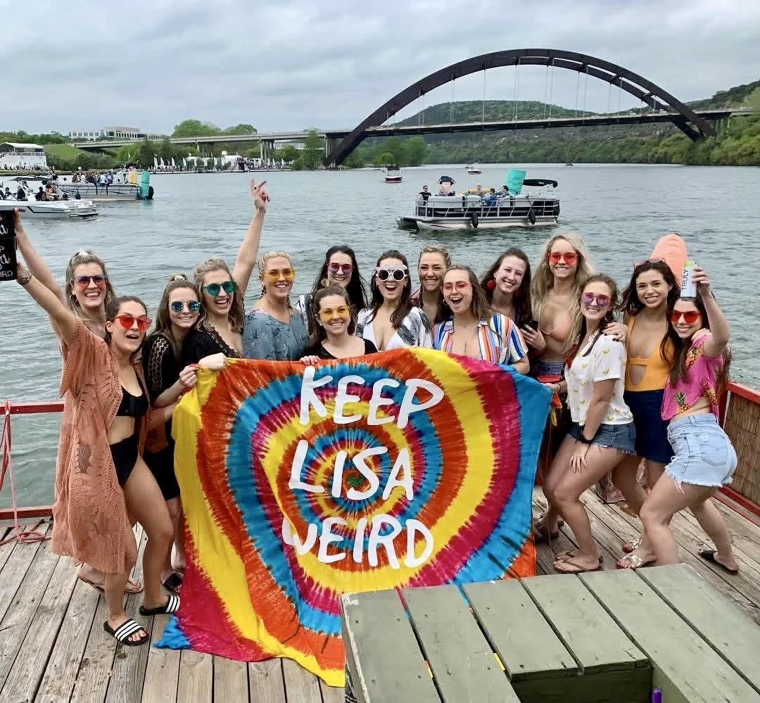 Group of women on a dock by a lake, holding a colorful tie-dye banner that reads 'Keep Lisa Weird,' with boats and a bridge in the background.
