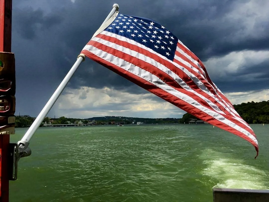 American flag waving on a boat with green water and a cloudy sky in the background.