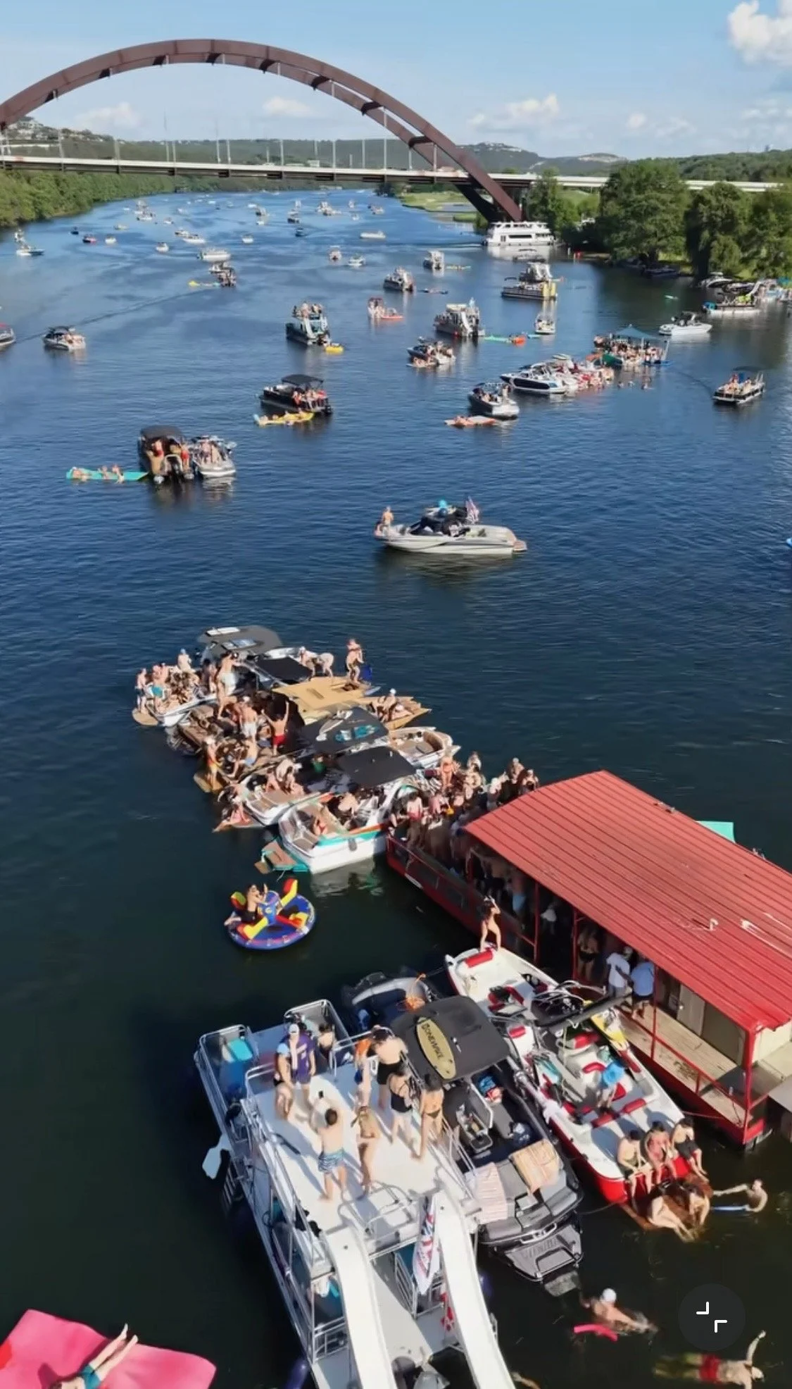 A crowded dock with boats and floating platforms filled with people enjoying a summer day near a river or lake. There is a bridge arched over the water in the background.