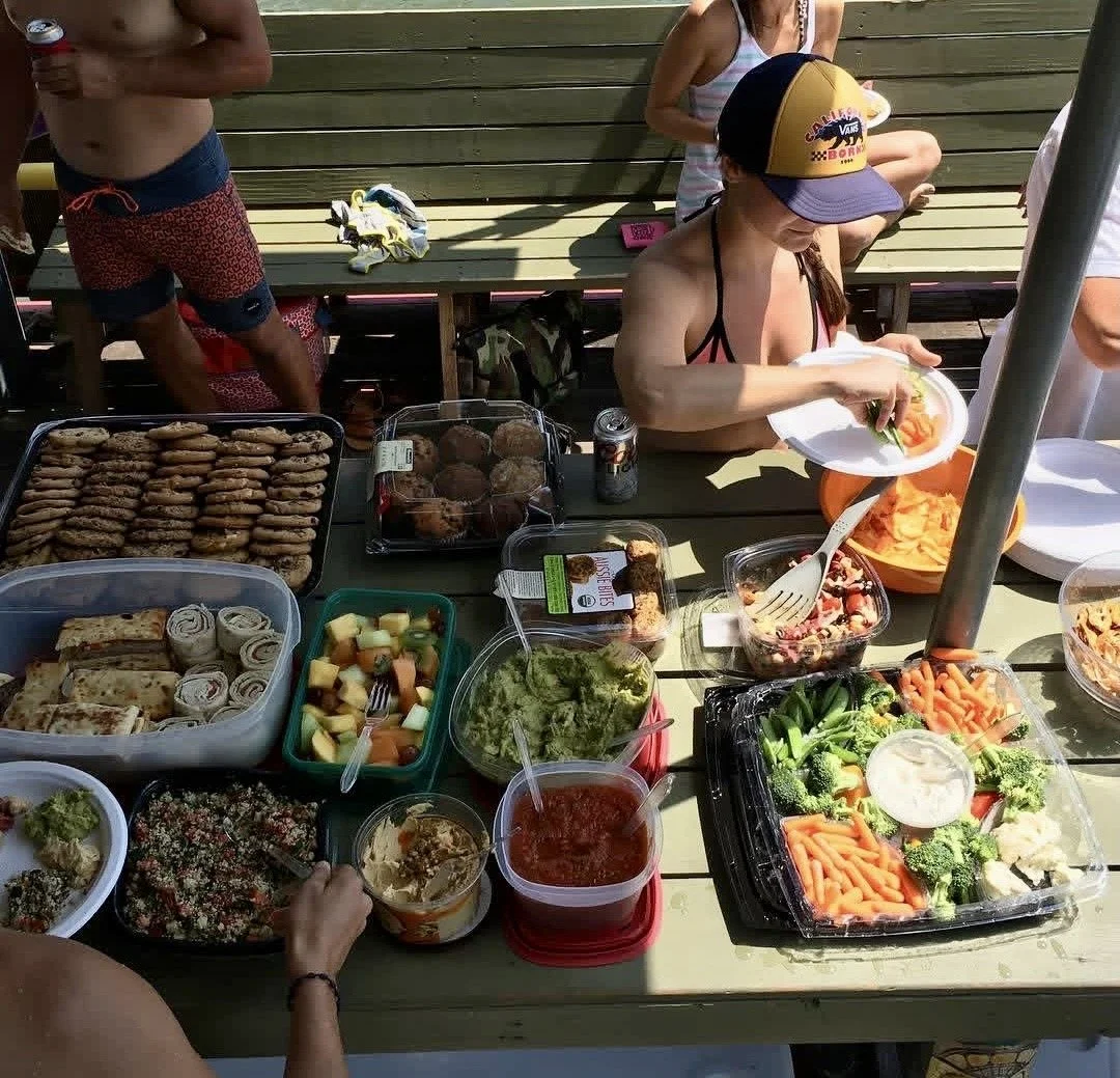 A table filled with various snacks and food items, including cookies, cupcakes, fruit salad, vegetables, and dips, with people around it enjoying a meal outdoors.