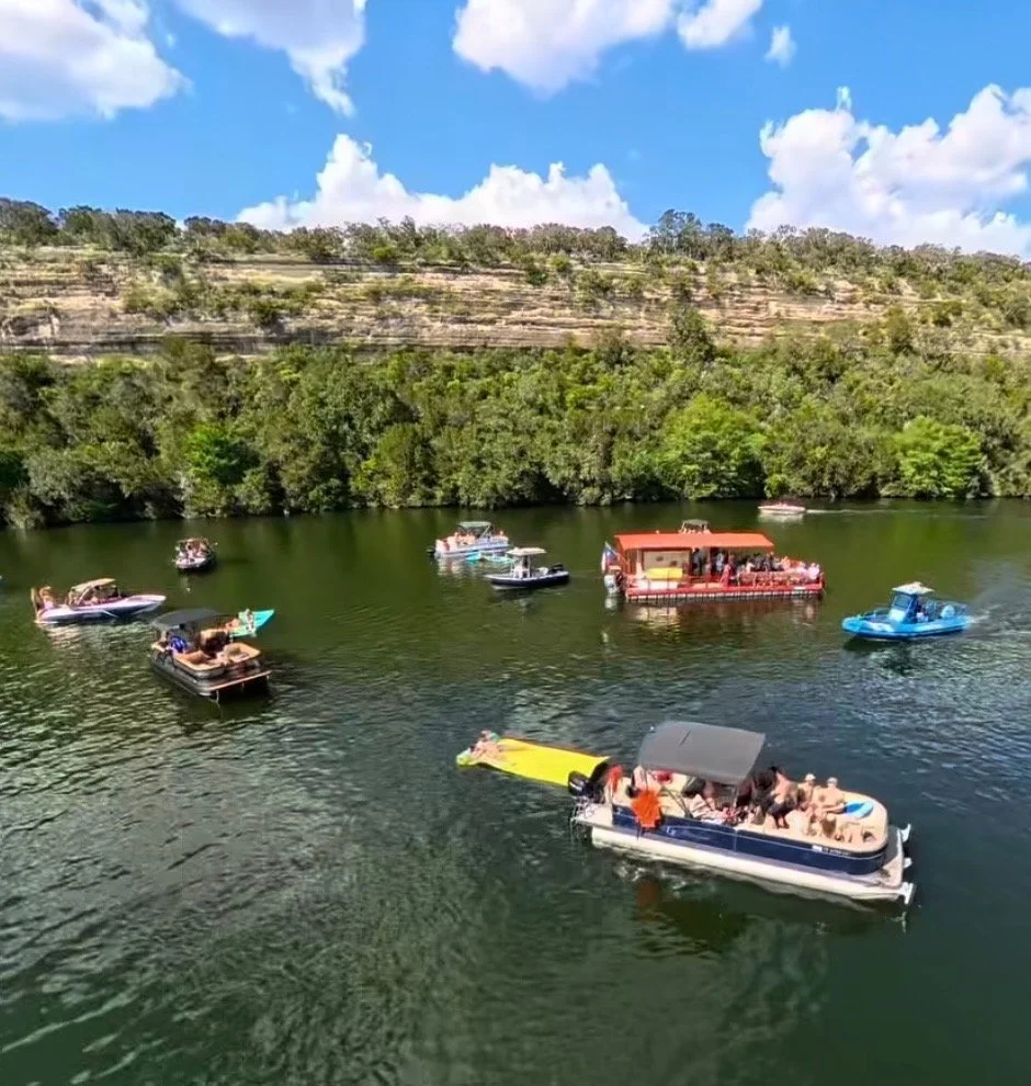 Multiple boats and floating platforms with people on a river, surrounded by lush green trees and rocky hills under a partly cloudy sky.