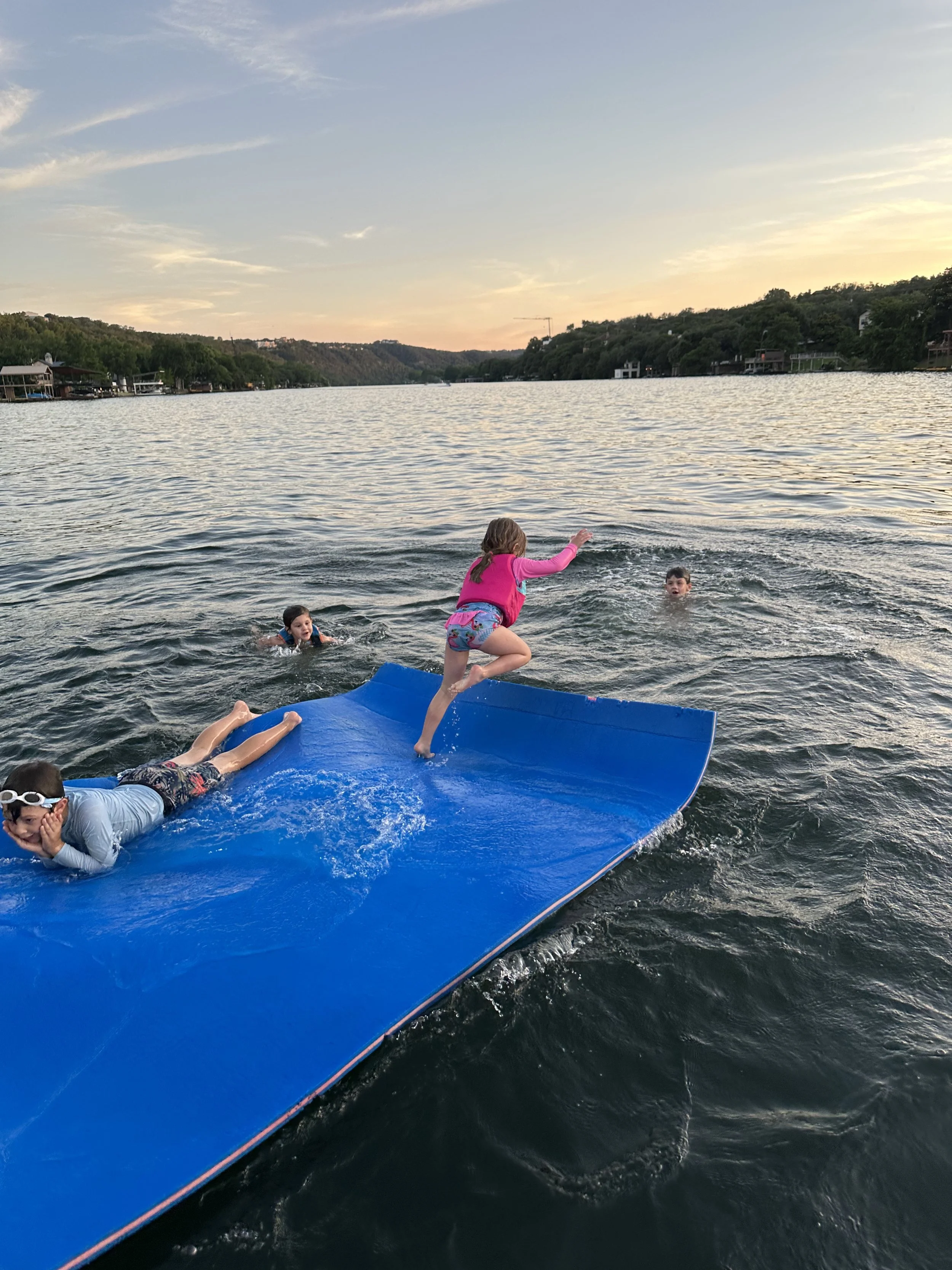 Children playing on a blue floating mat in a lake at sunset, with hills and houses in the background.