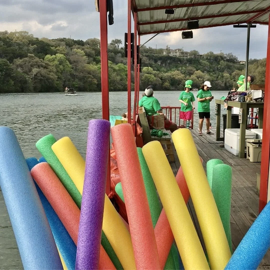 Colorful pool noodles in the foreground on a boat with people standing and sitting on a dock by a lake with dense trees and hills in the background.