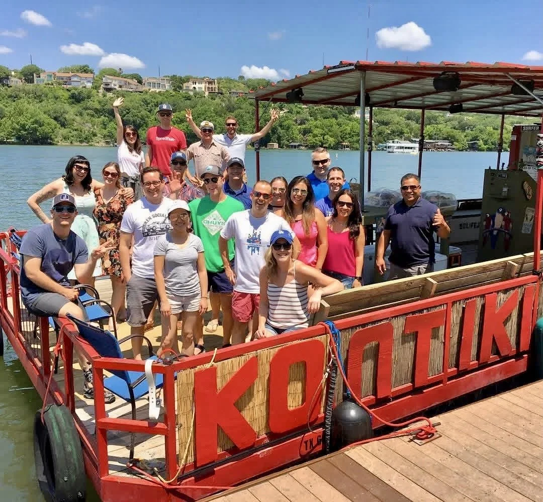 Group of people on a boat named 'KOLUKI' on a river with green hills and houses in the background under a blue sky with some clouds.