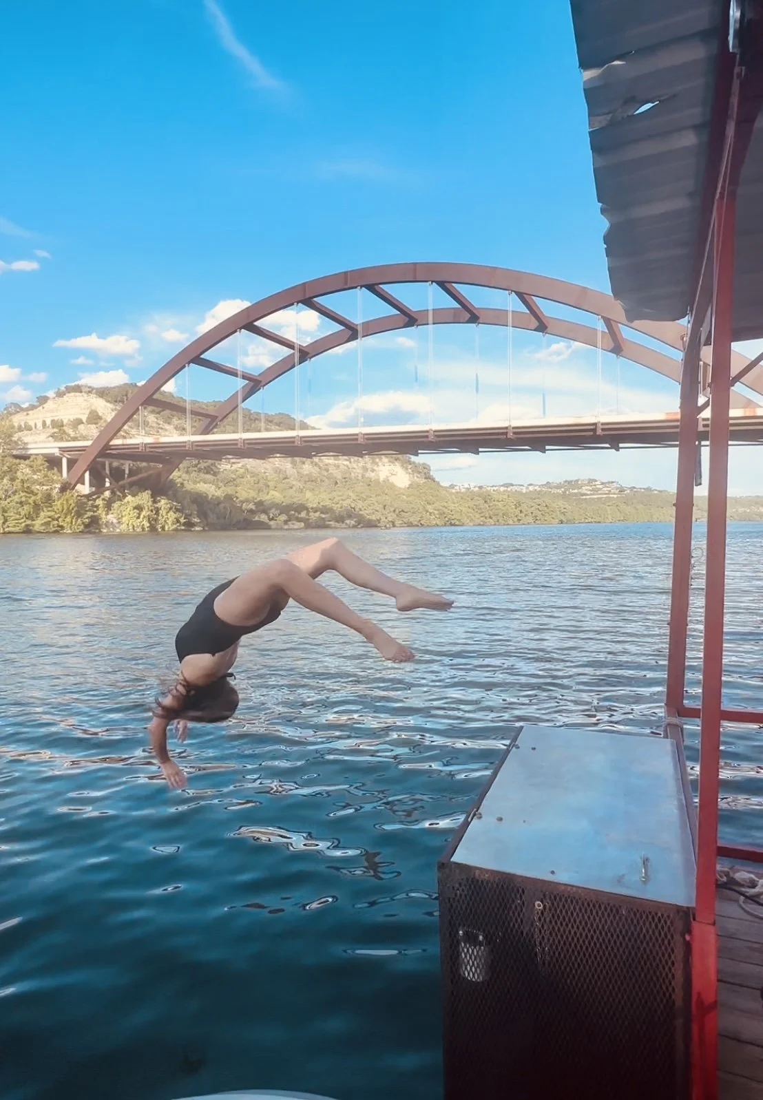 A person in a swimsuit performing a backflip off a dock into a body of water, with a bridge and hills in the background on a clear day.