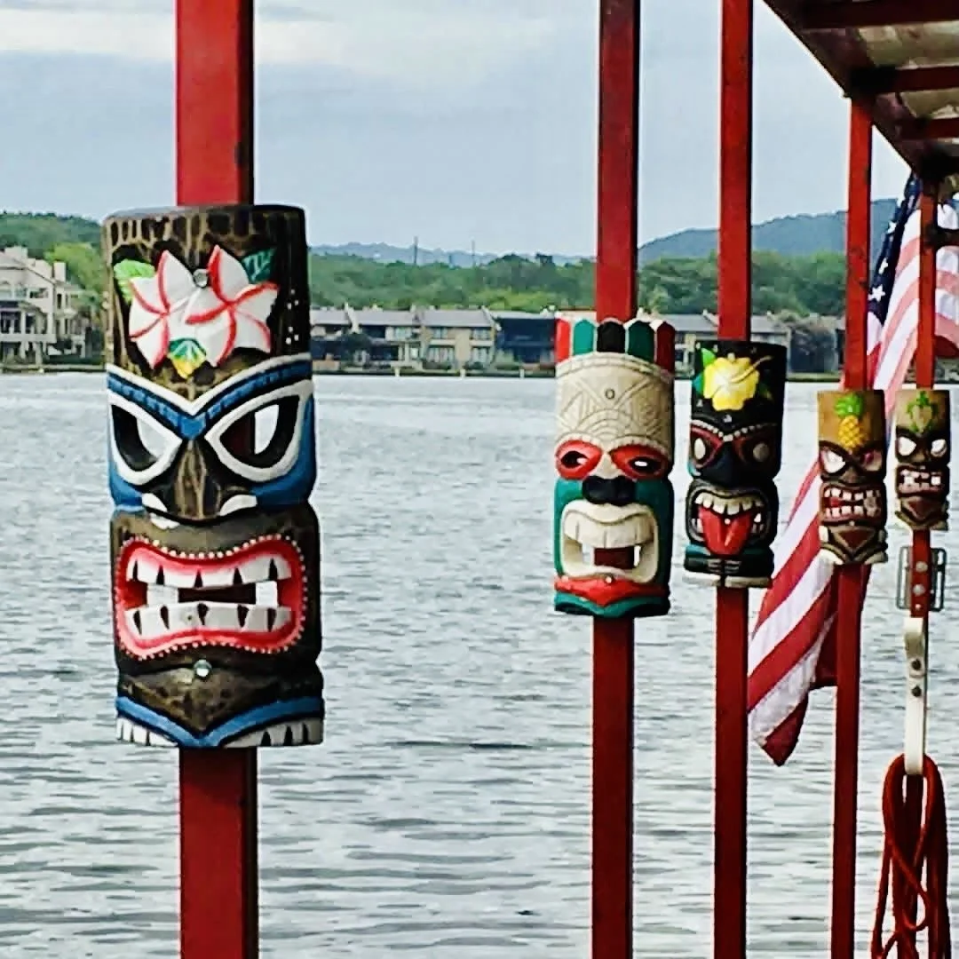 Colorful wooden tiki masks hanging on red poles over water, with houses and hills in the background.
