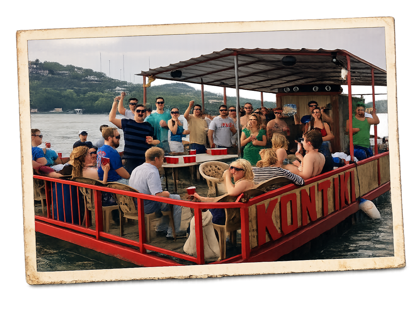 A group of people celebrating on a floating boat named 'KONTIKI' on a river during daytime, with some standing and raising drinks, others sitting and socializing, surrounded by scenic greenery and hills in the background.