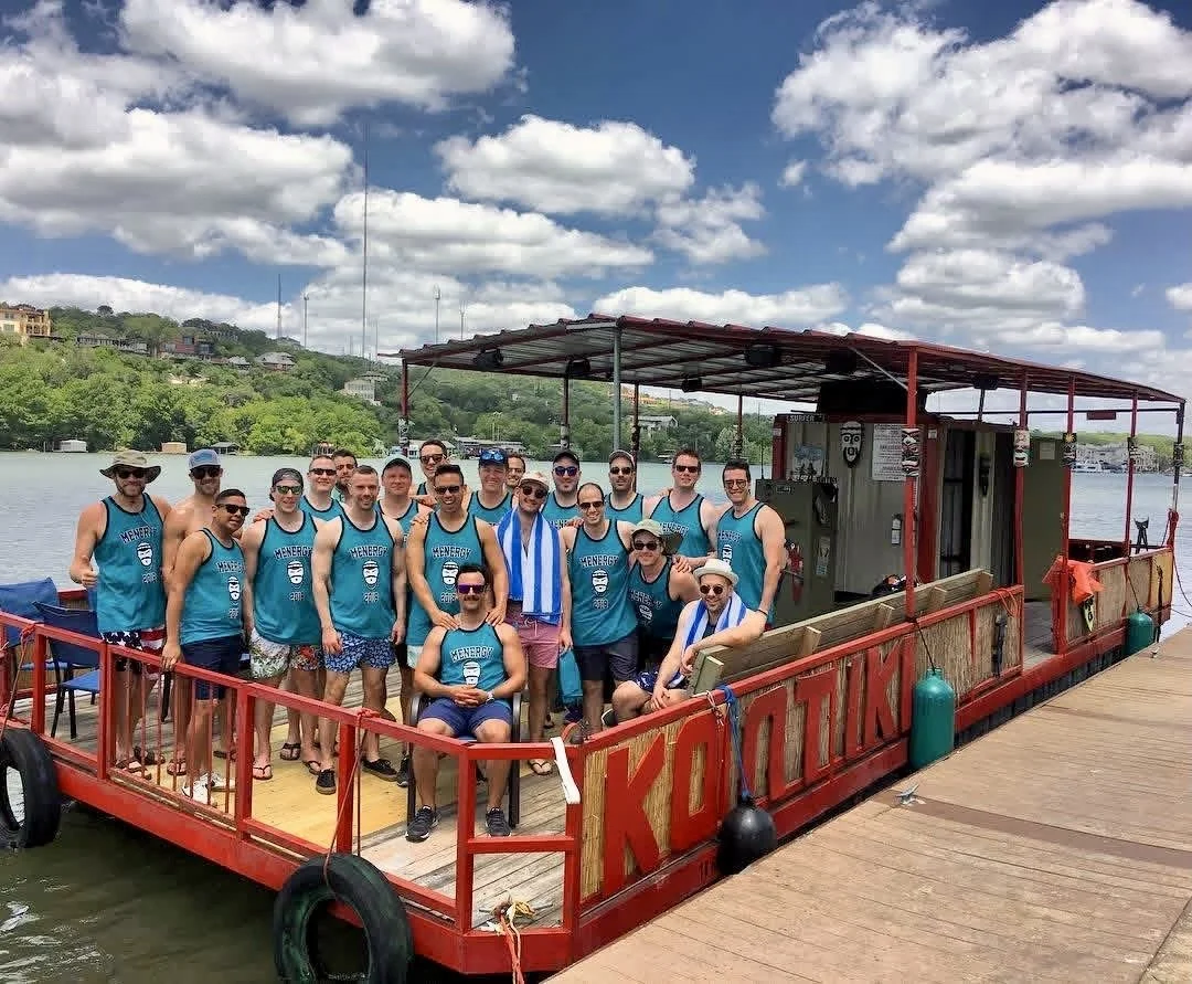 Group of people on a boat docked on a river, wearing summer clothing and life jackets, with a background of water, trees, and a partly cloudy sky.