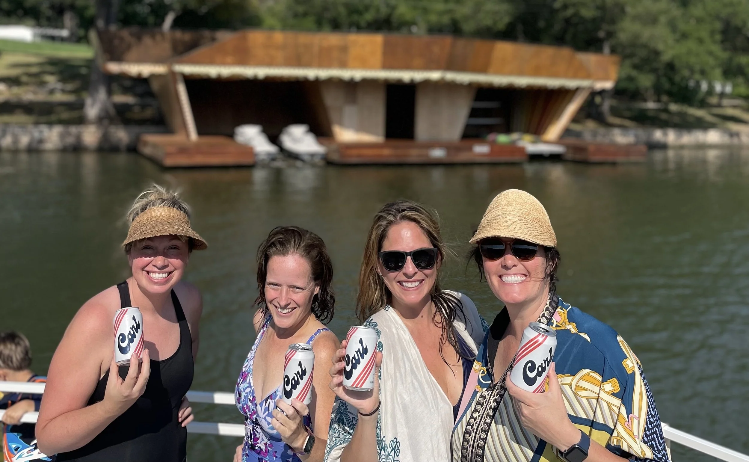 Four women smiling and holding cans of Coors beer in front of a body of water with a modern, boat house in the background, during daytime.