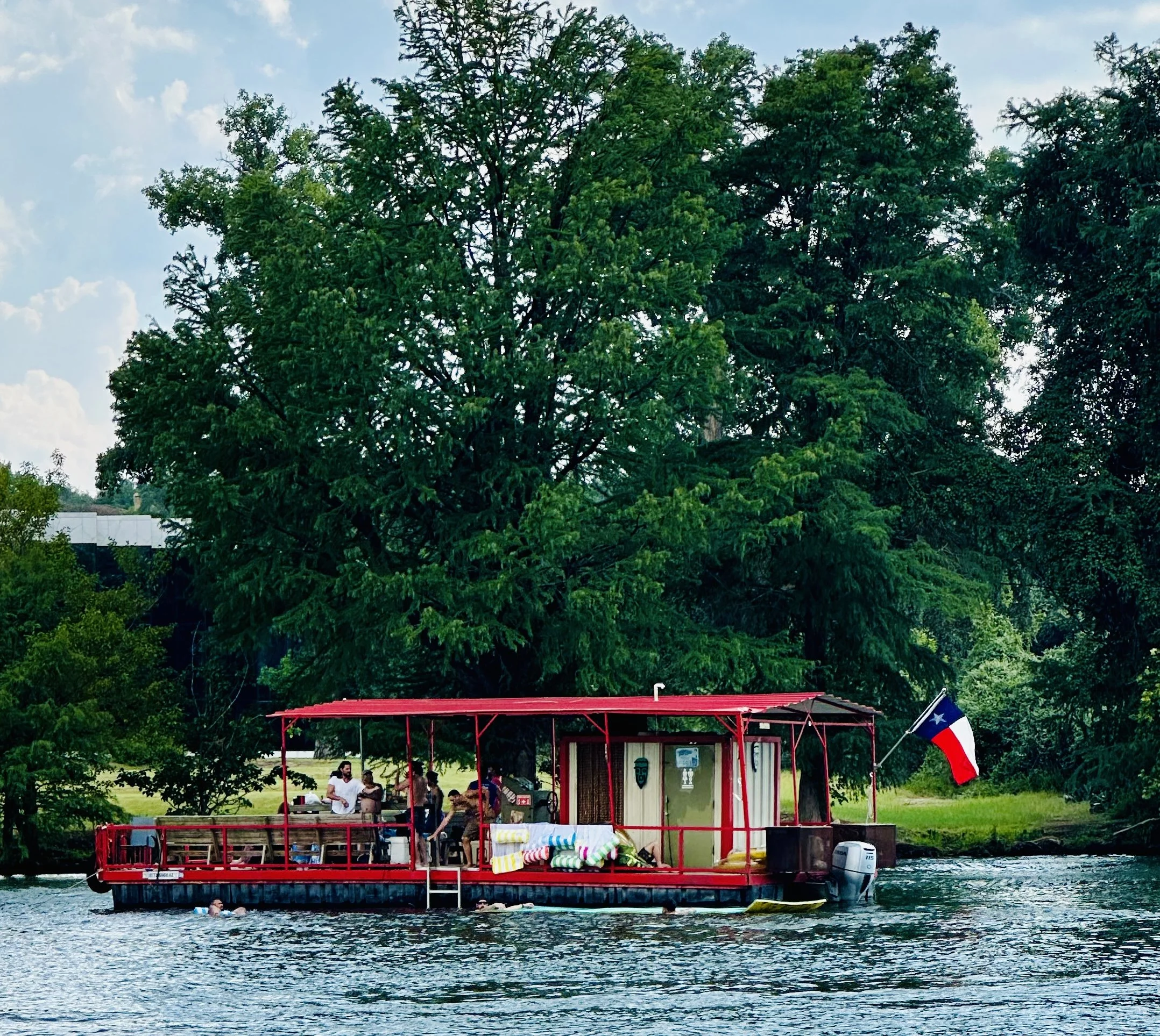 A houseboat on a body of water with several people gathered on it. The houseboat has a red roof structure and a Texas flag at the corner. Large trees and a blue sky with clouds are in the background.
